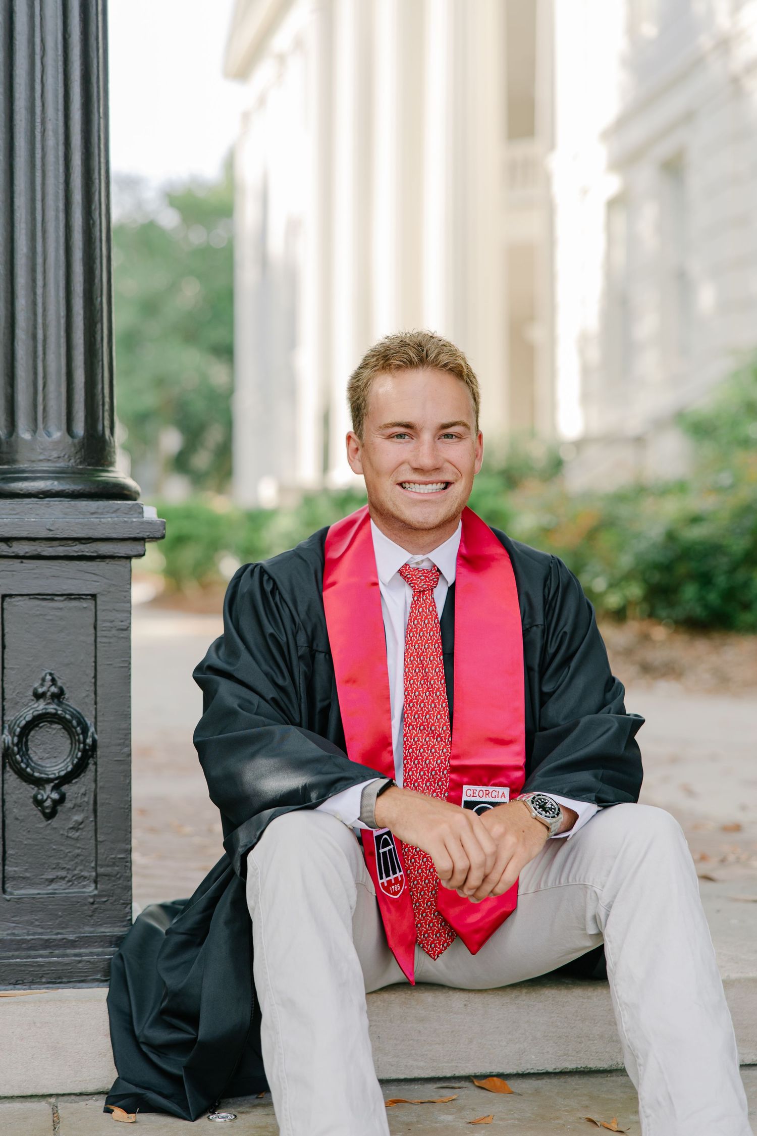 Male graduate in black gown with red stole sitting on steps smiling.