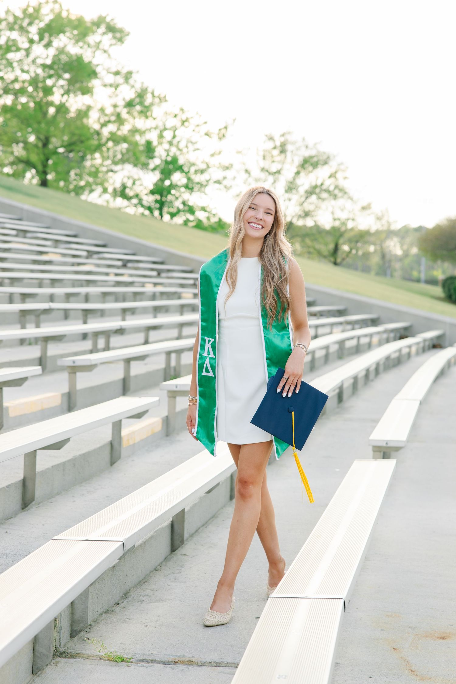Smiling female graduate in white dress with Kappa Delta green stole walking down stadium bleachers.