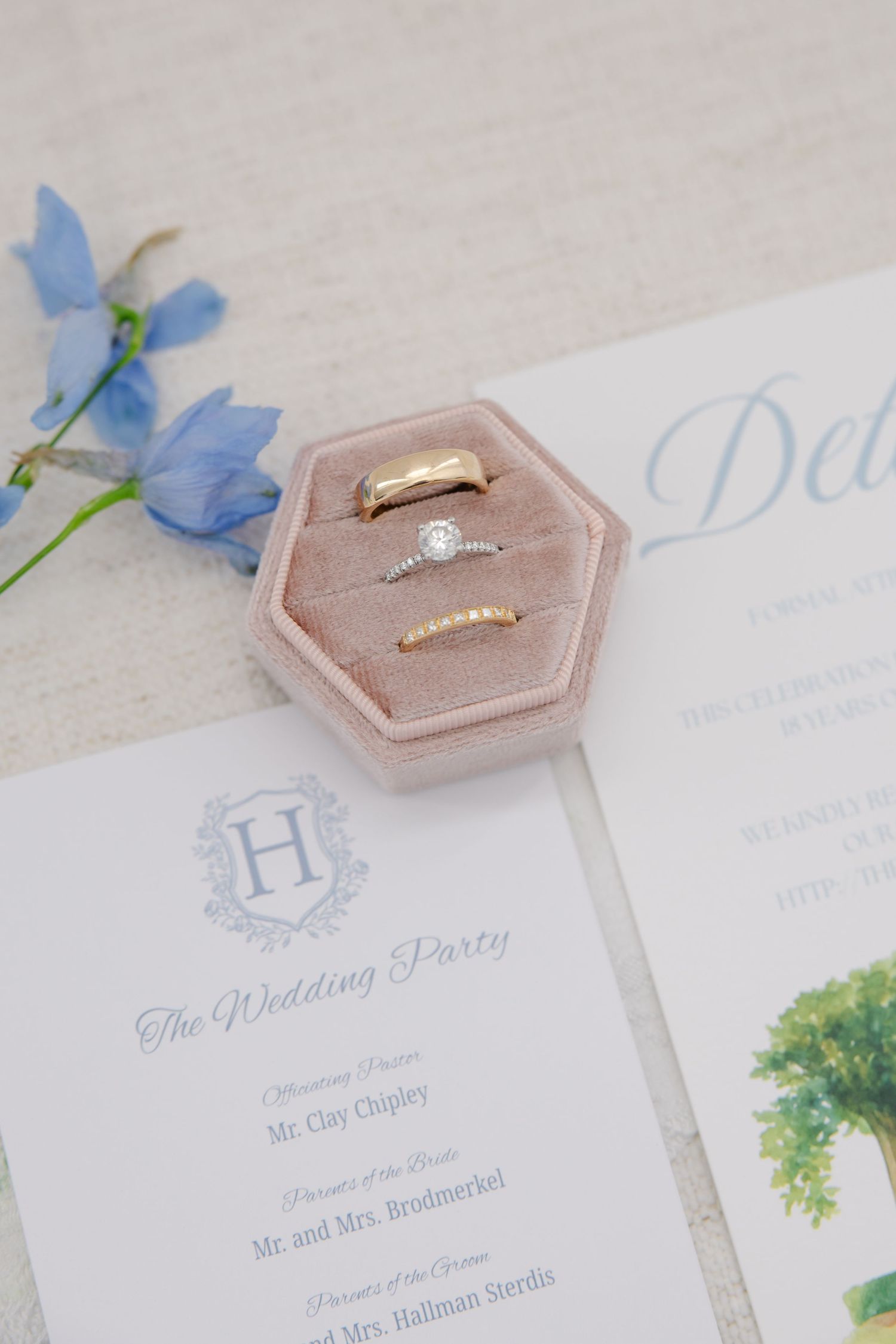 Wedding rings displayed in pink velvet hexagon box alongside floral invitation suite and blue flowers.