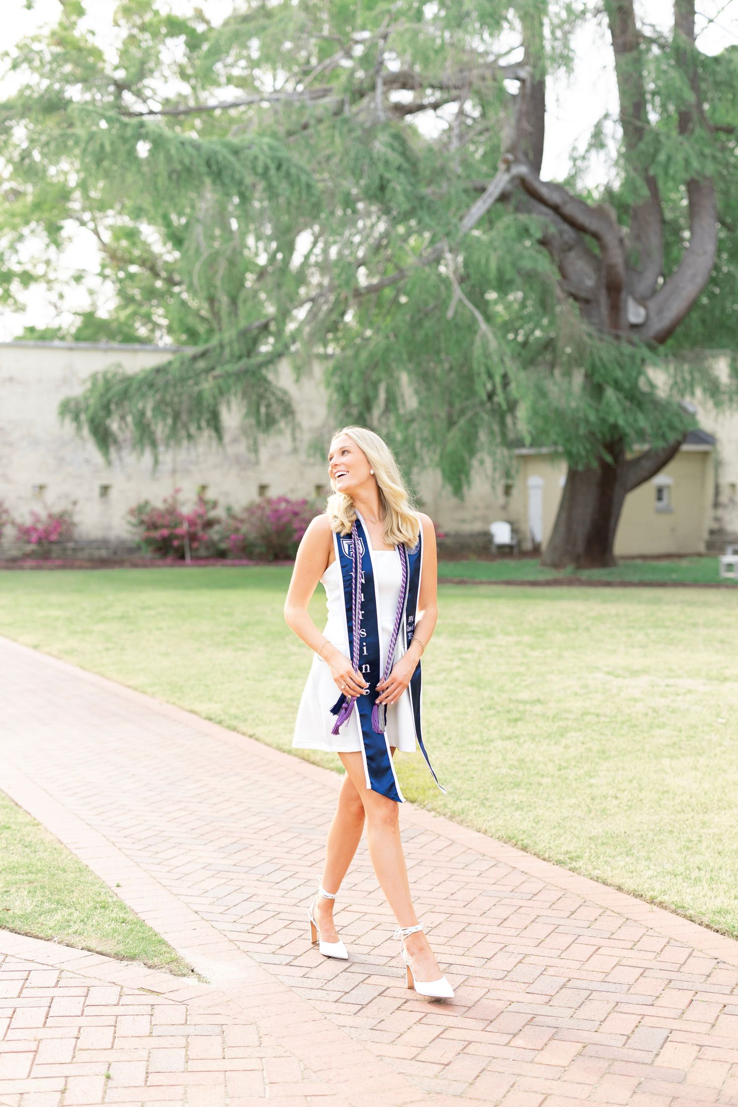 Nursing graduate in white dress and navy graduation stole walks sunny brick campus pathway.