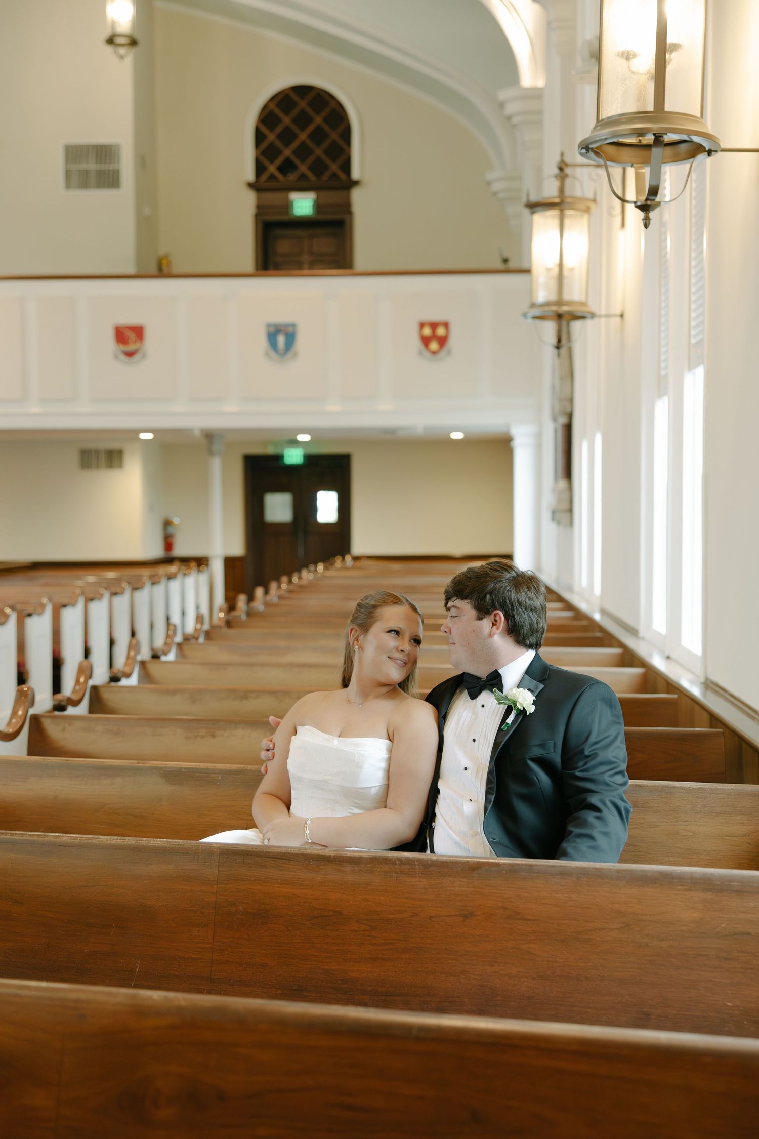Bride and groom sit closely together in wooden church pews, sharing a tender moment on their wedding day.