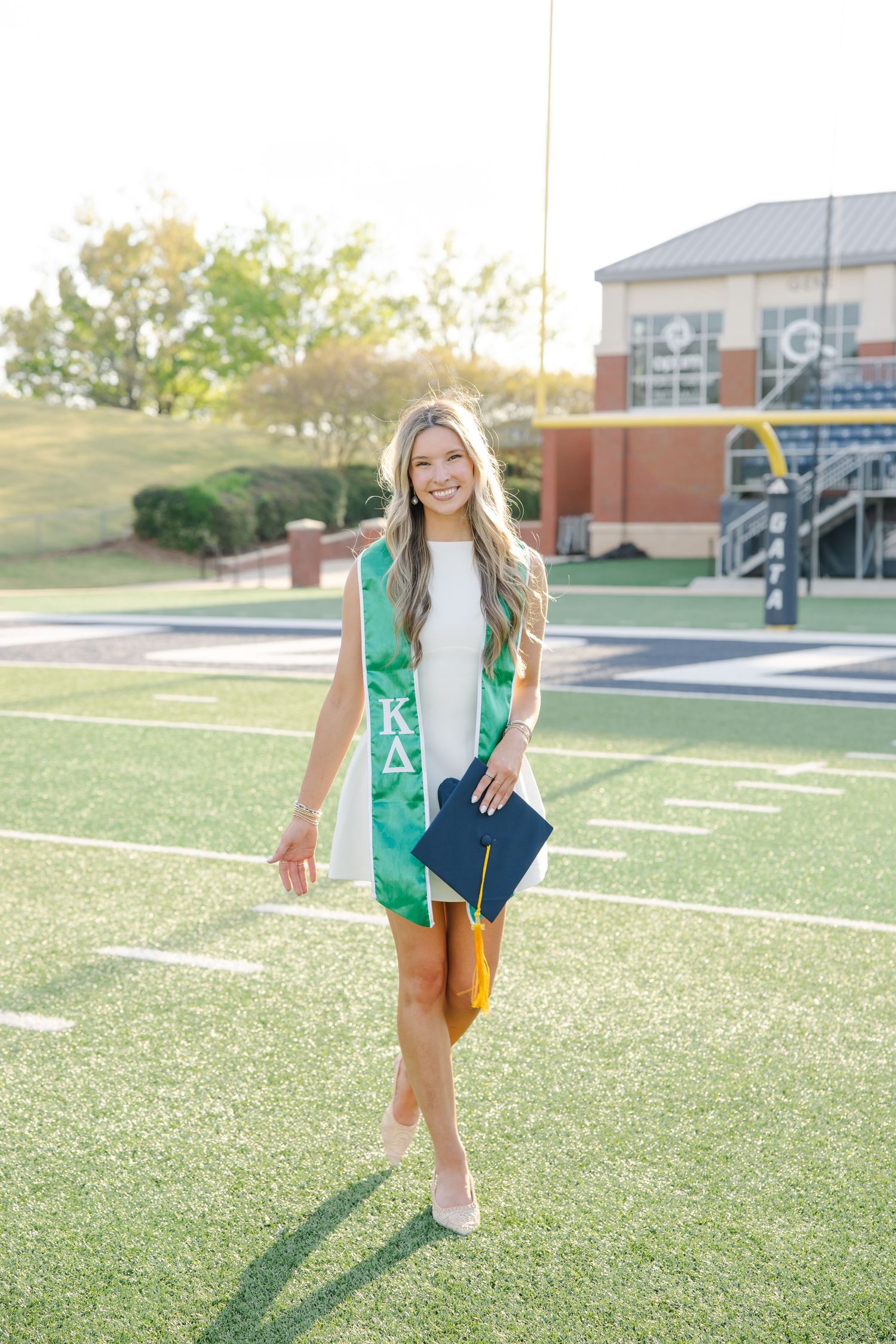 Joyful female graduate in white dress with Kappa Delta stole walking across college football field.