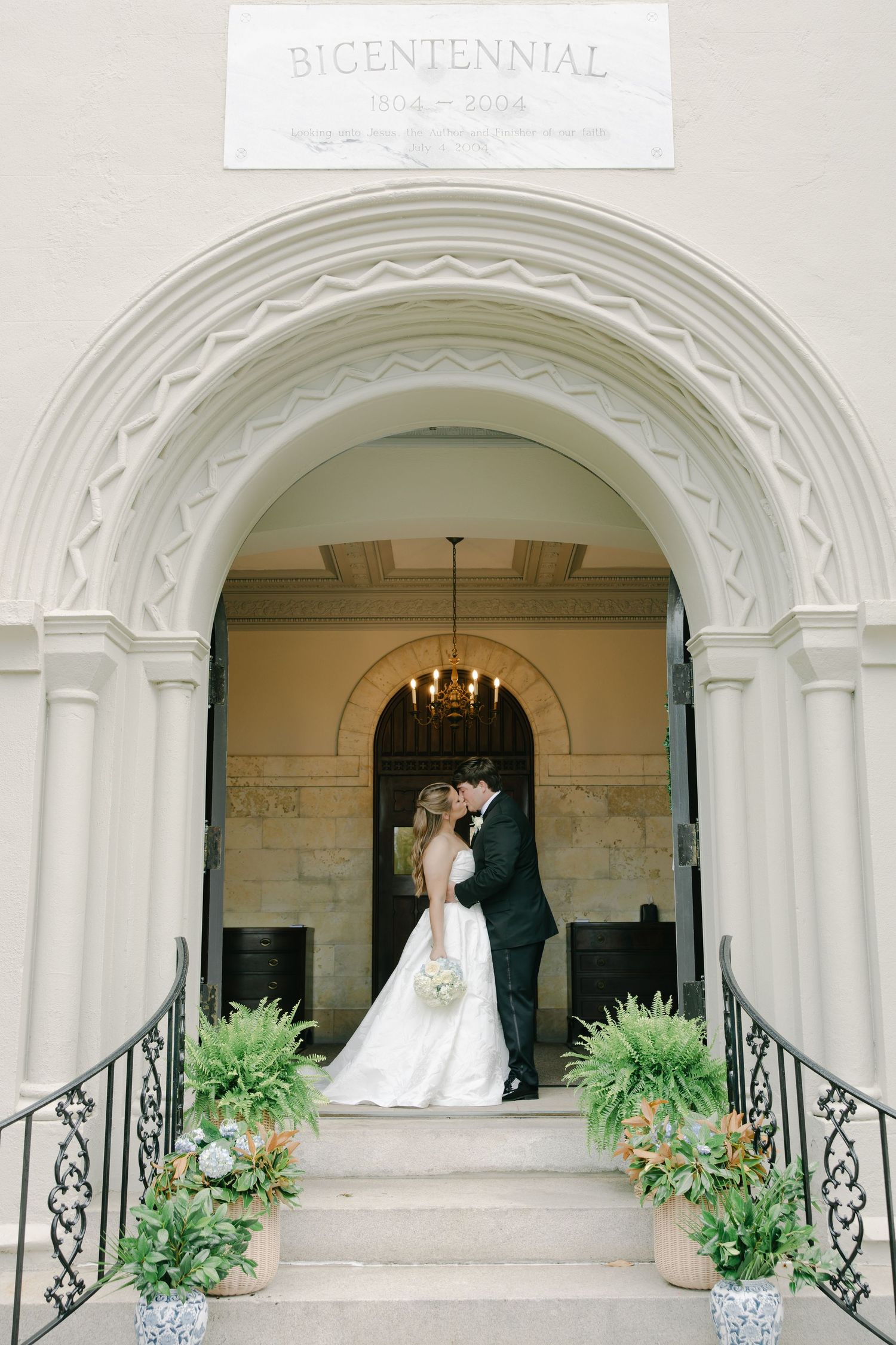 Bride and groom share a kiss under an ornate arched doorway of a historic Bicentennial building adorned with lush greenery.