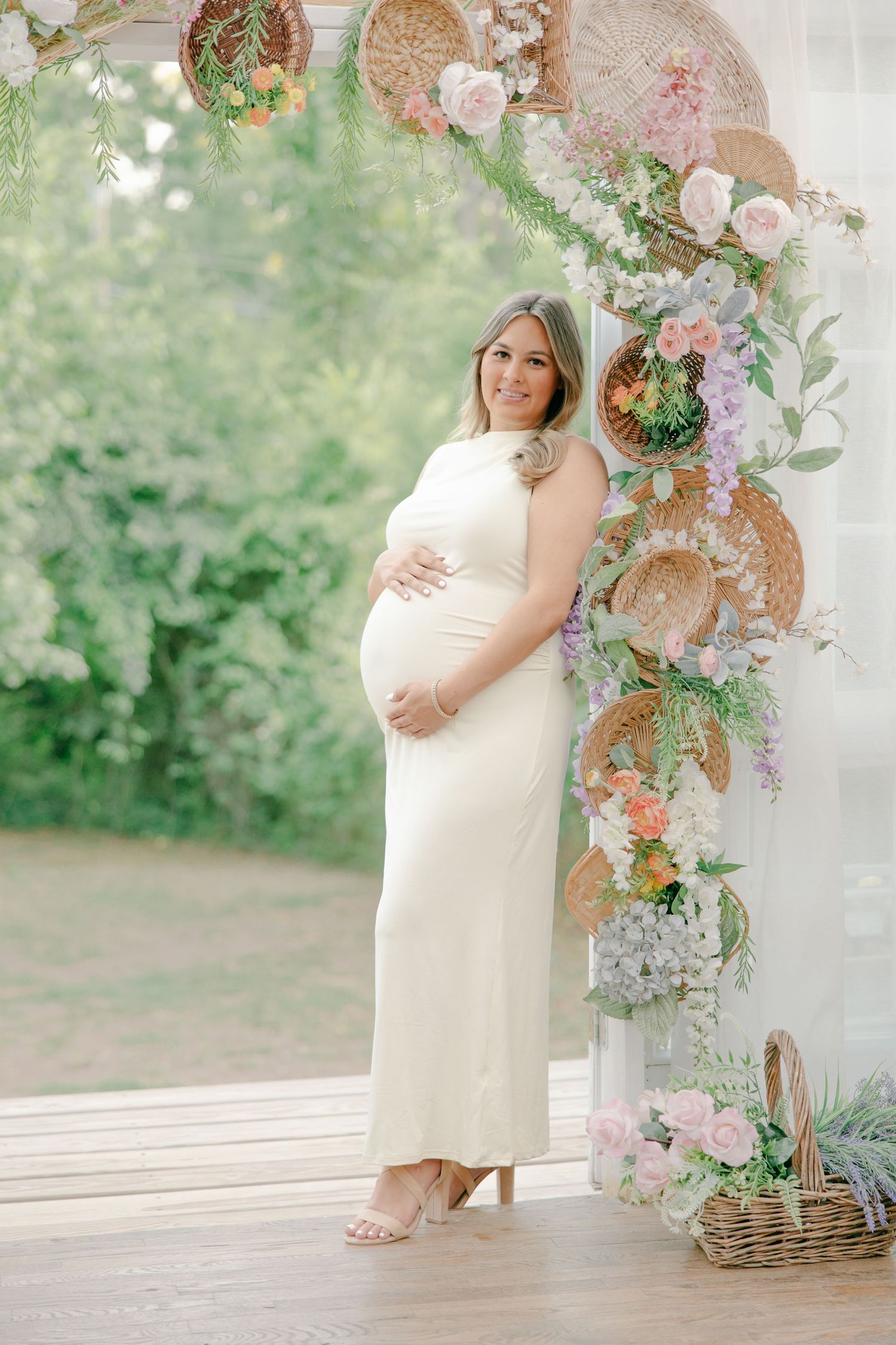 Beautiful pregnant woman in white dress poses beside a boho floral arch with wicker accents outdoors.