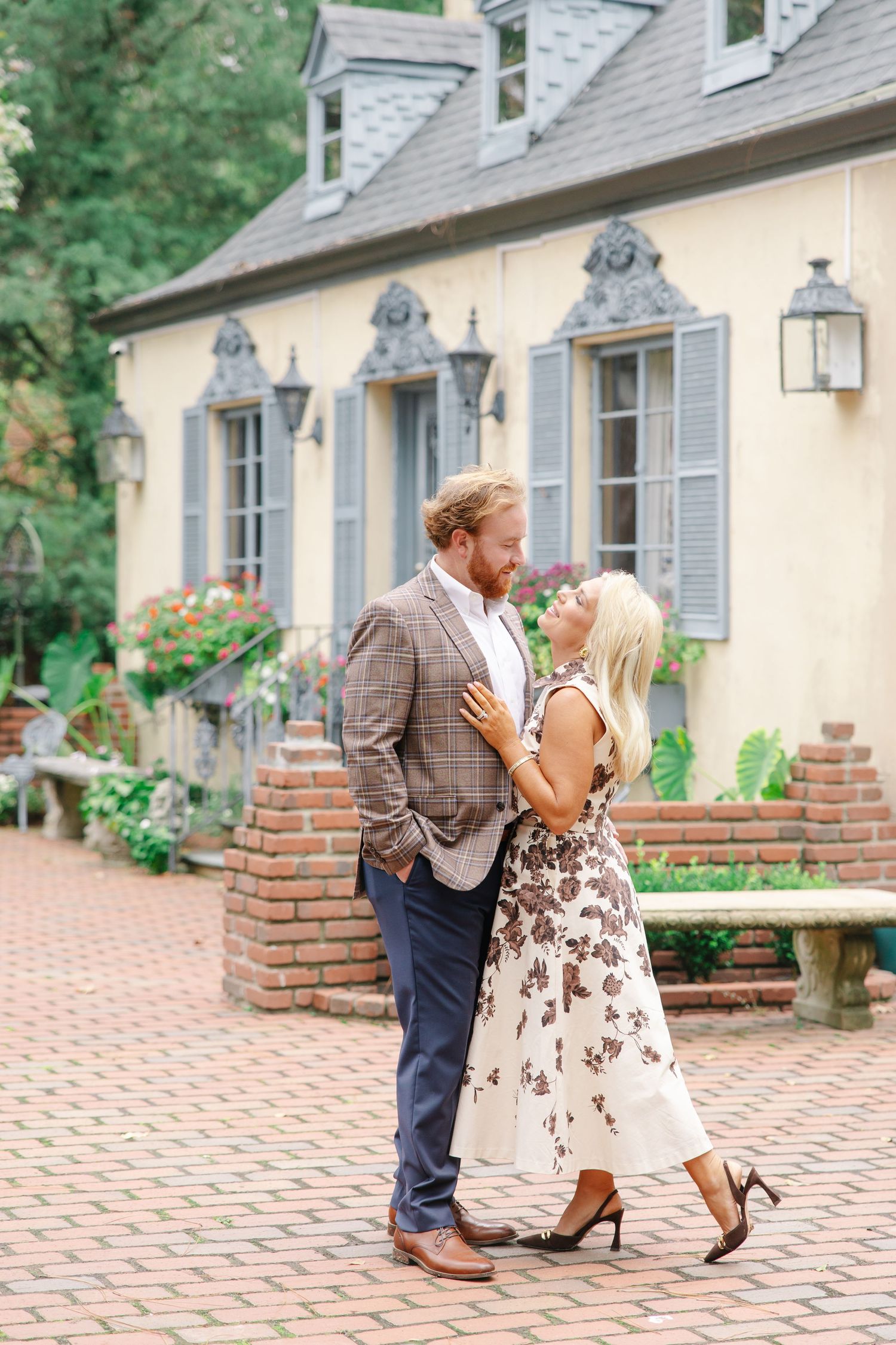 Couple sharing a tender moment on brick courtyard in front of charming yellow French-style estate.