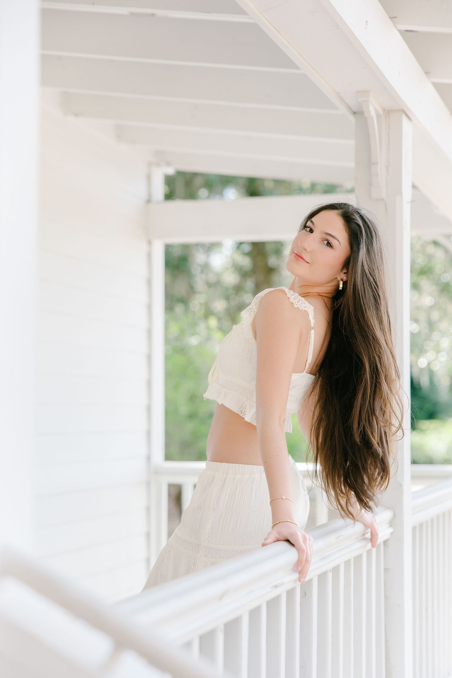 Young woman with long dark hair in cream lace crop top and skirt leaning on white porch rail.
