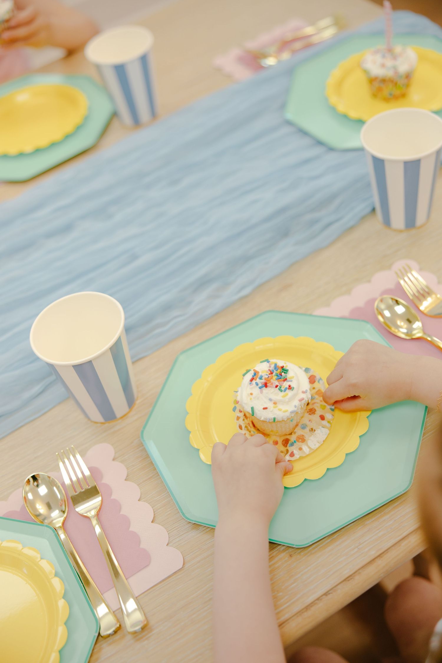 Child's hands placing a cupcake with colorful sprinkles on a pastel party table setting with gold cutlery.