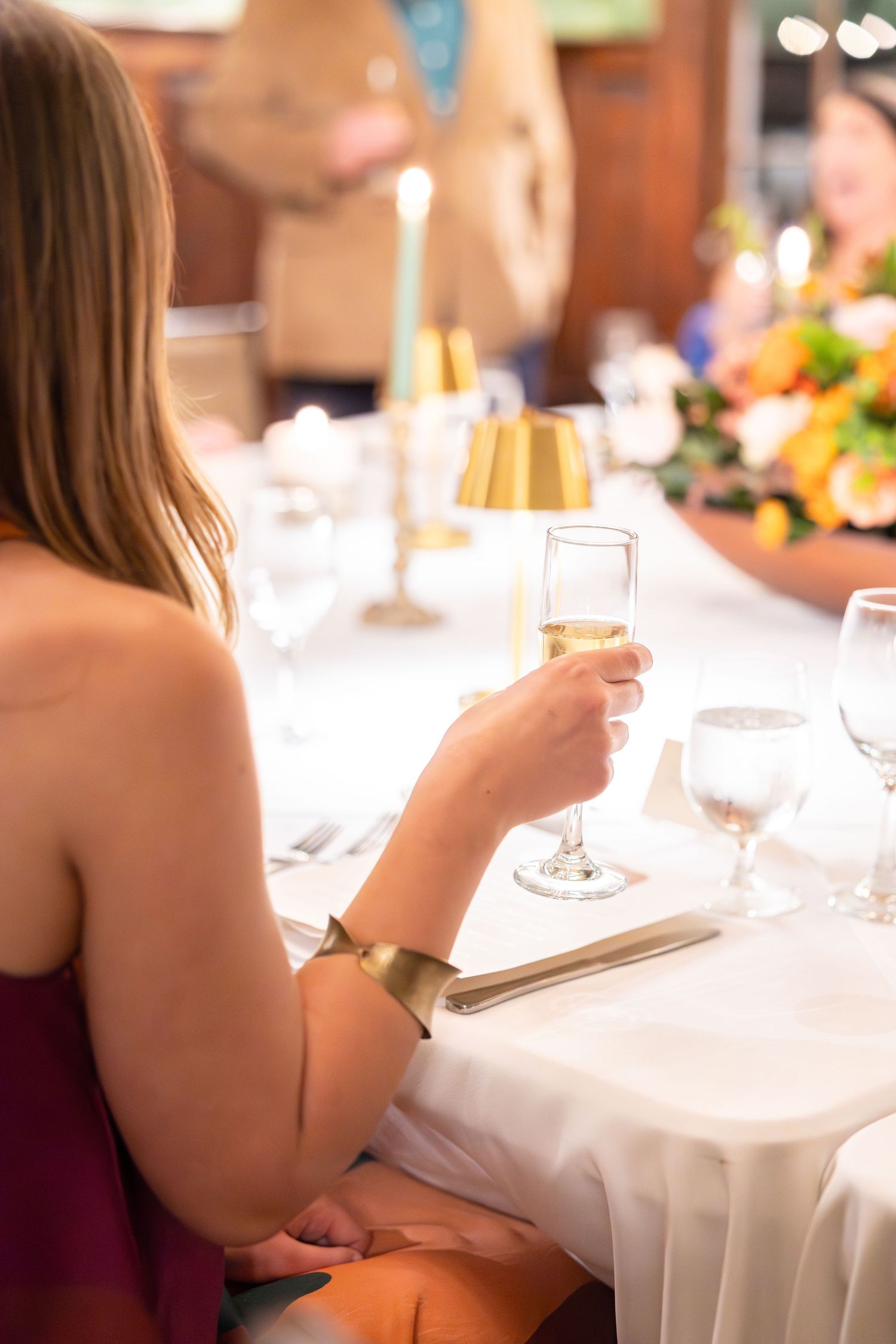 Woman in burgundy dress holding champagne glass at elegantly set dinner table with gold accents and floral centerpiece.