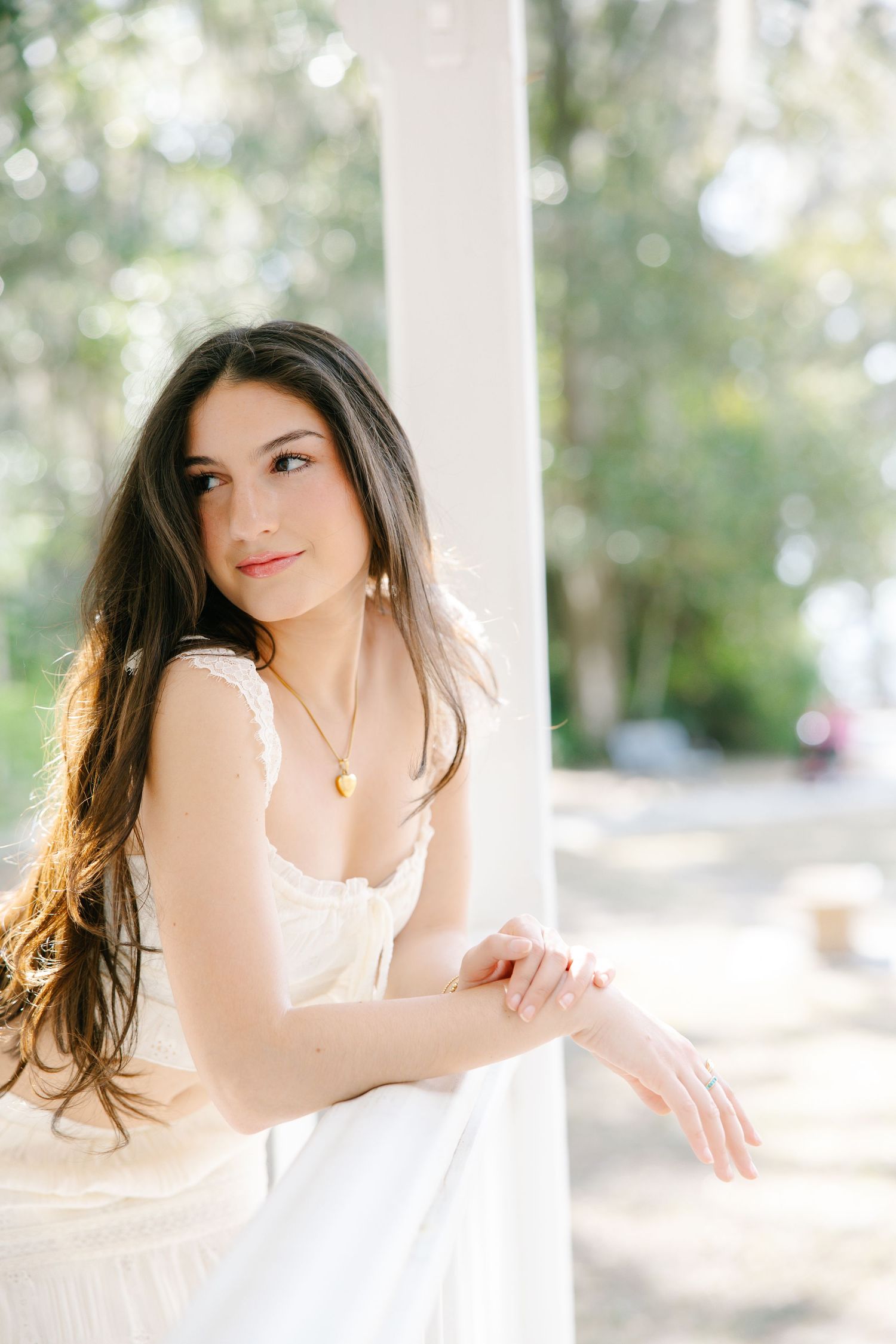 Beautiful brunette teen in white dress leans on white column in soft natural outdoor light.