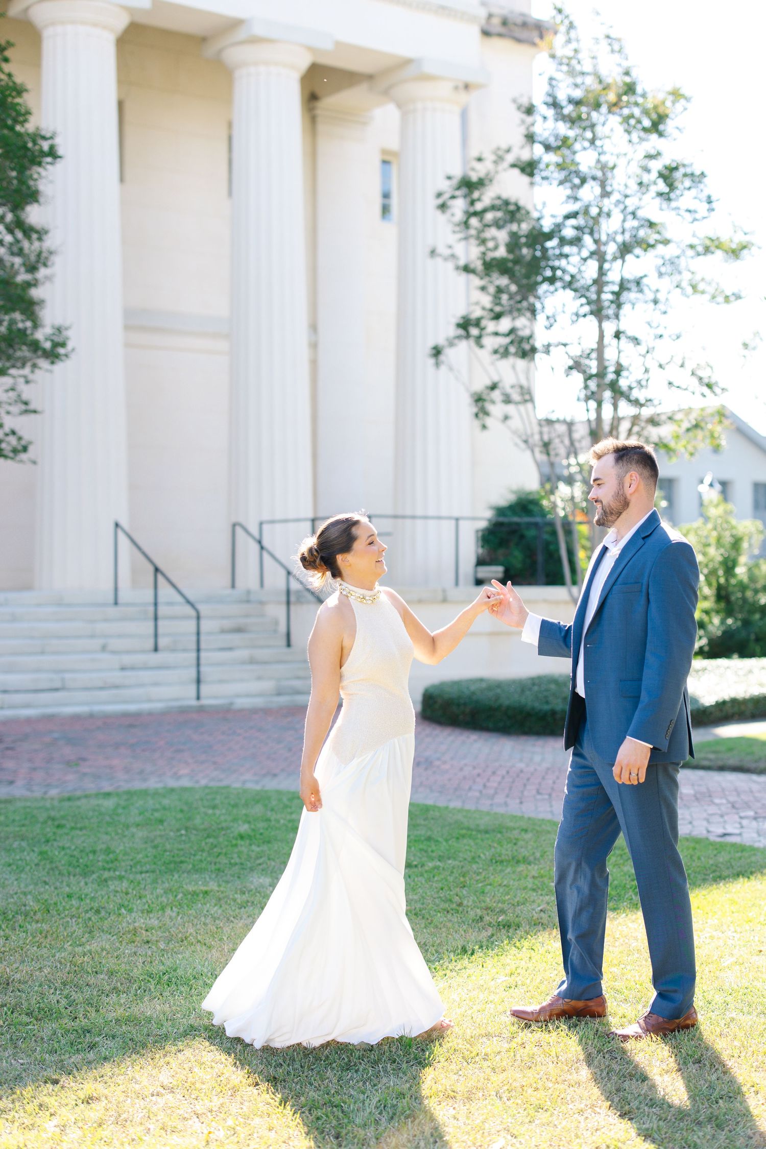 Bride in elegant white gown and groom in blue suit dancing on a sunlit lawn.