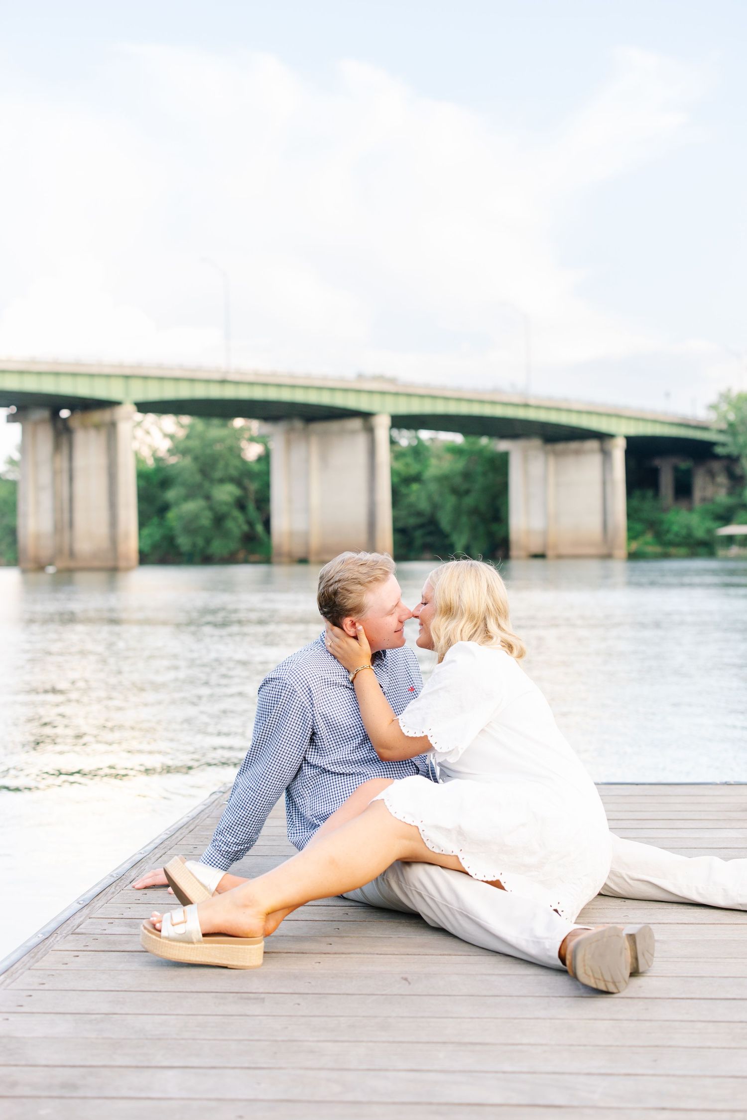 Couple sharing a kiss while sitting on wooden waterfront dock with bridge behind them.