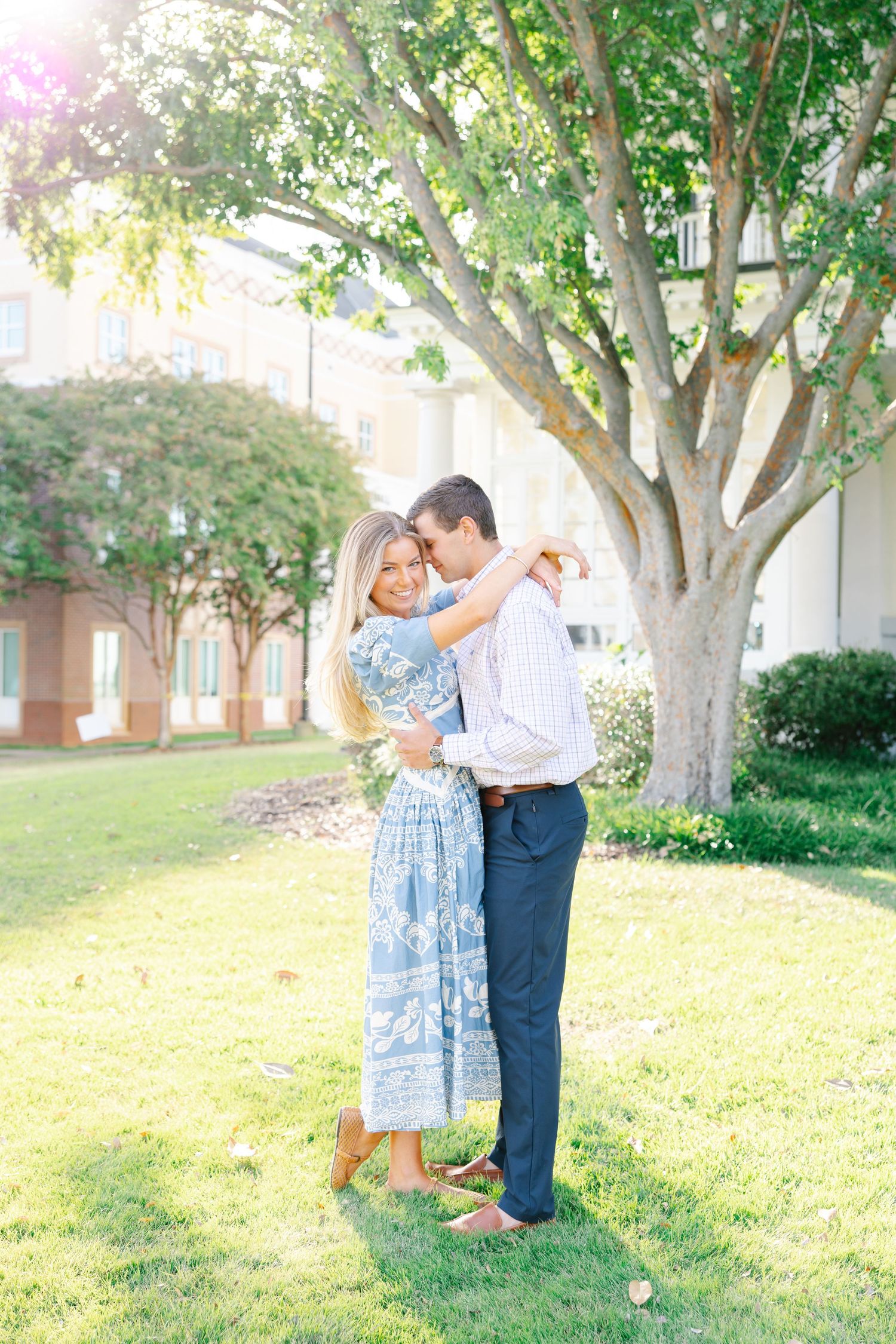 Smiling blonde woman in blue floral dress embracing man under large tree on sunny lawn.