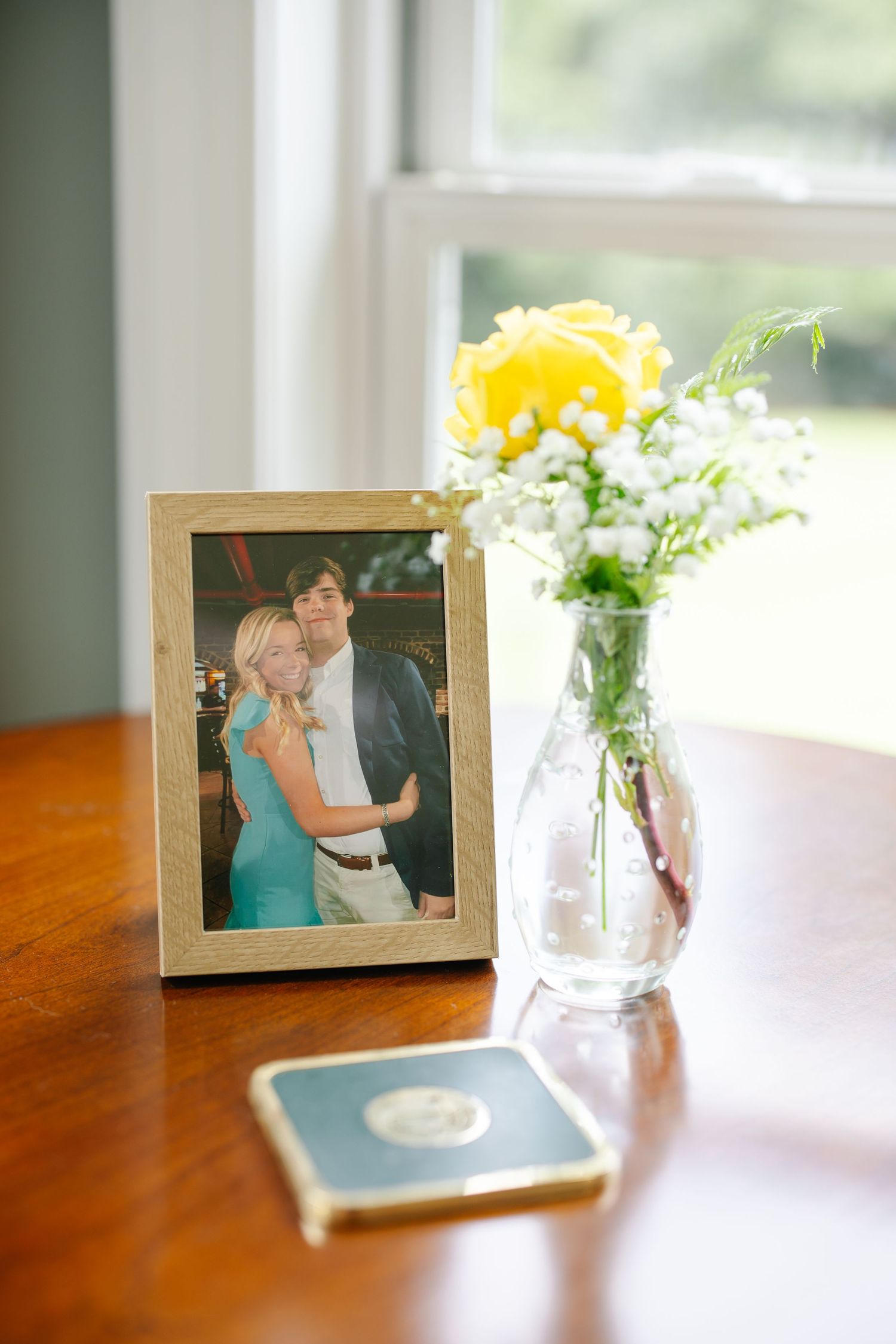 Framed couple photo on wooden table beside yellow rose and baby's breath in glass vase by window.