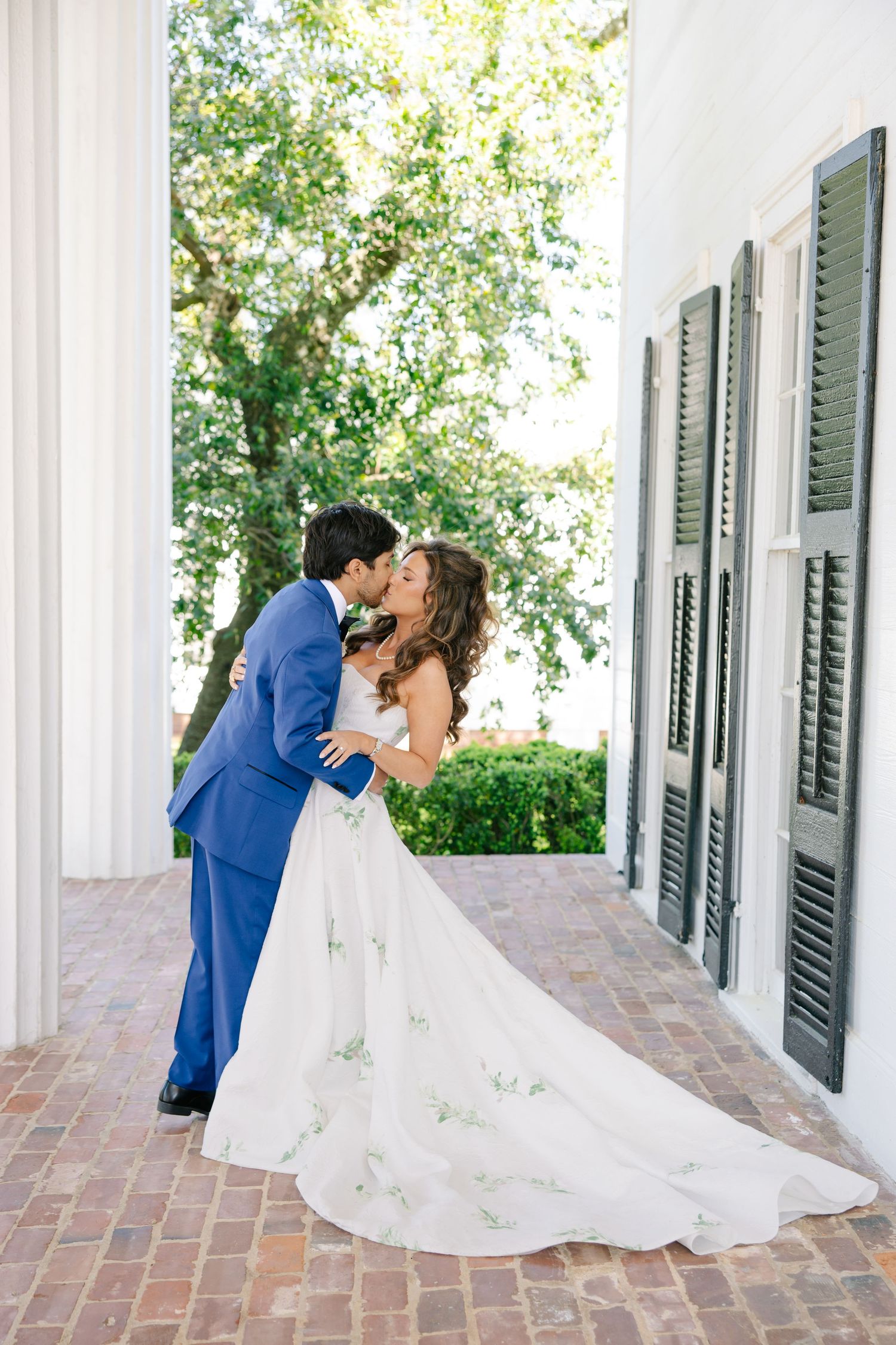 Bride and groom share a passionate kiss beside white columns, her floral gown train spread across brick porch floor.