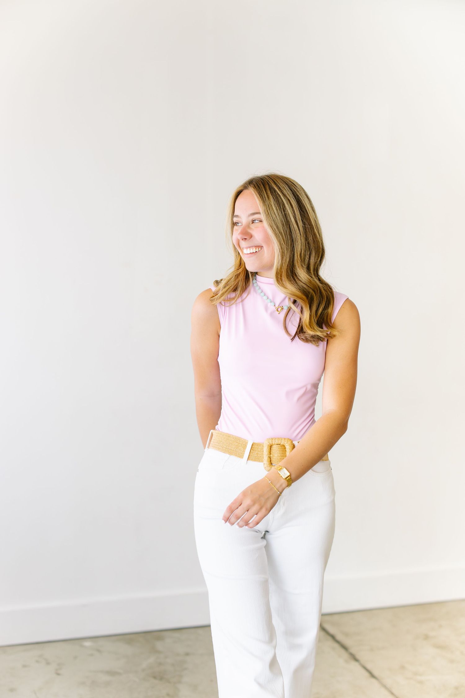 Happy woman in pink top and white pants with gold belt looking to the side against a white wall.
