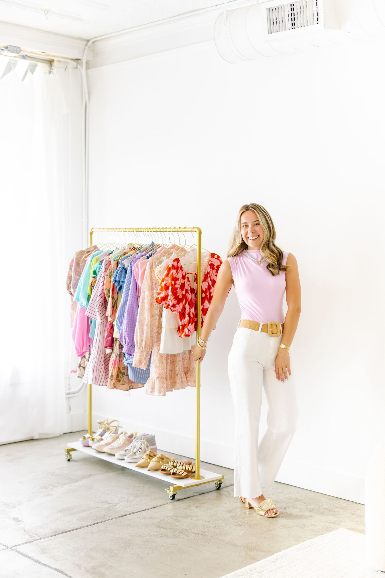 Smiling woman in pink top and white pants poses beside a gold clothing rack filled with colorful garments.