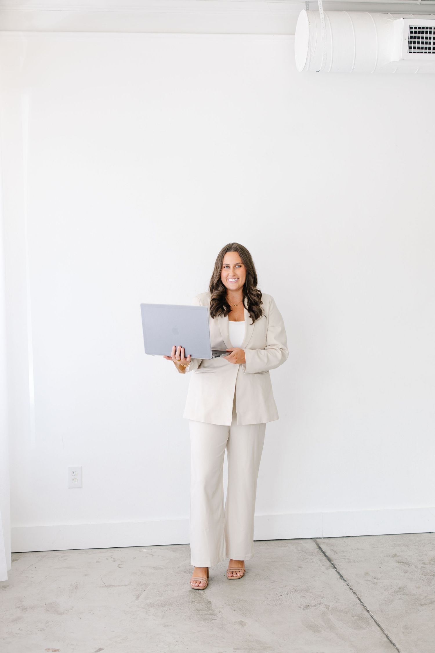 Professional brunette woman in beige pantsuit holds an open laptop in a bright white studio space.