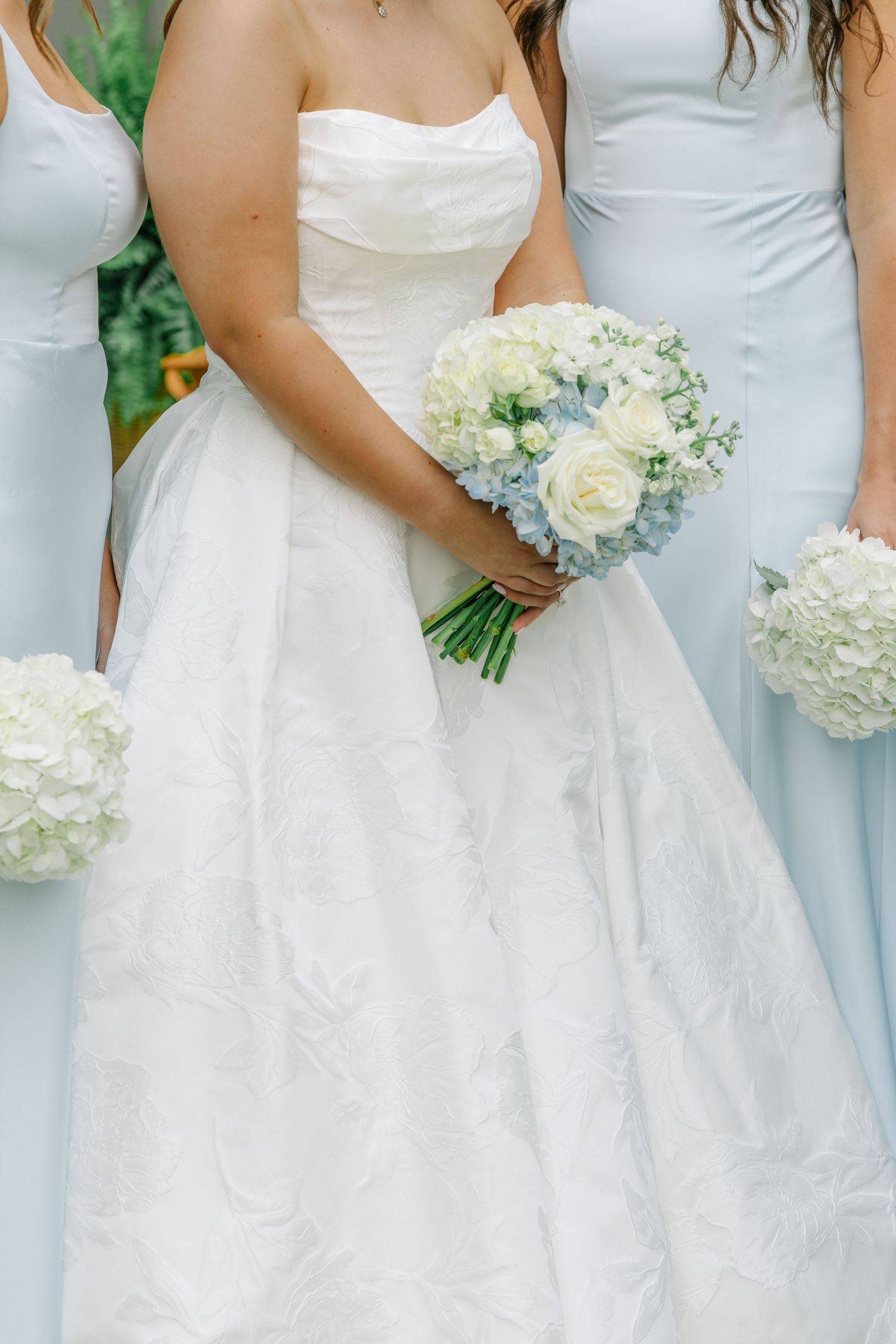 Bride in strapless white gown holds bouquet of white roses and blue hydrangeas beside bridesmaids in light blue dresses.