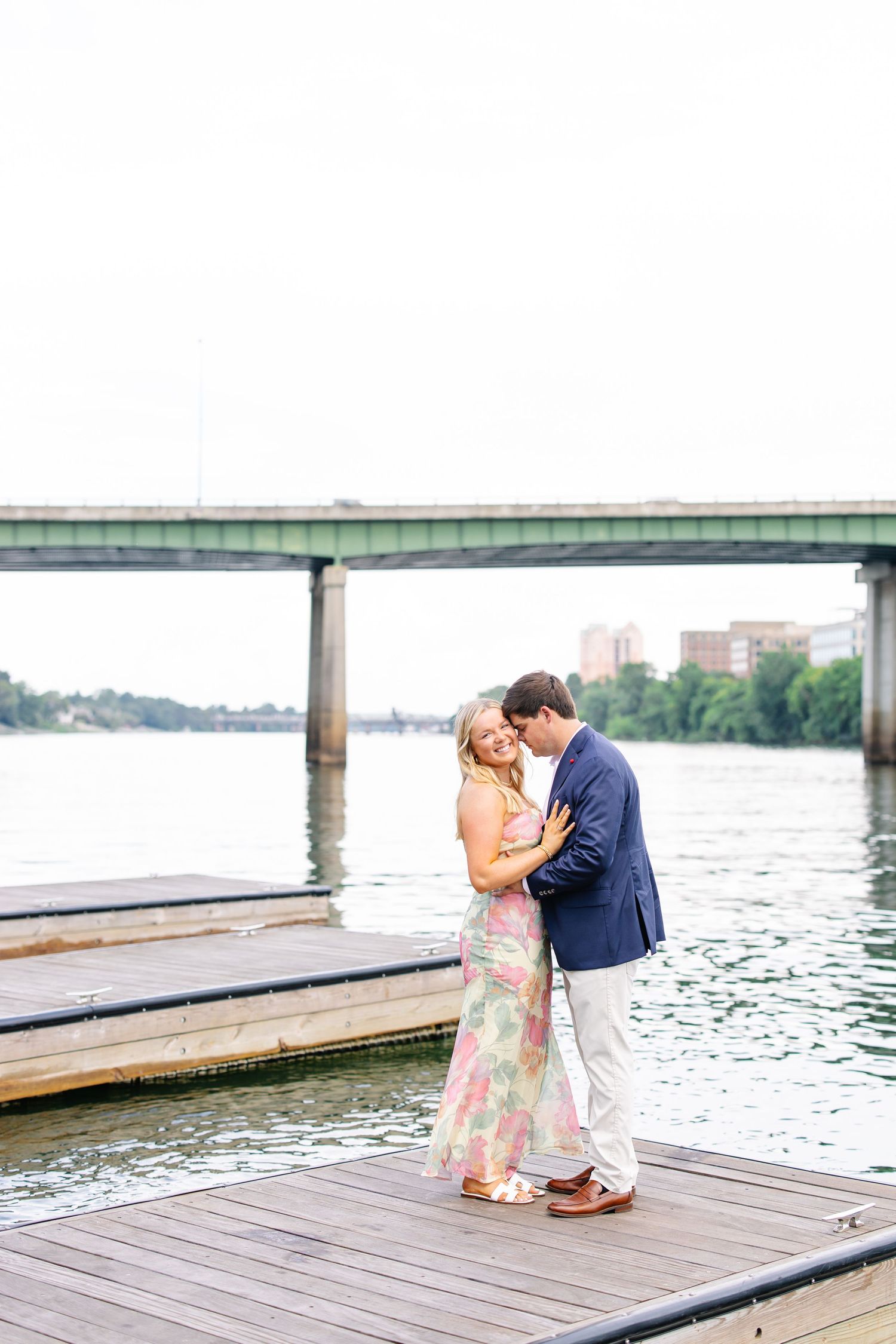 Newly engaged couple embracing on waterfront dock with bridge and city skyline in background.