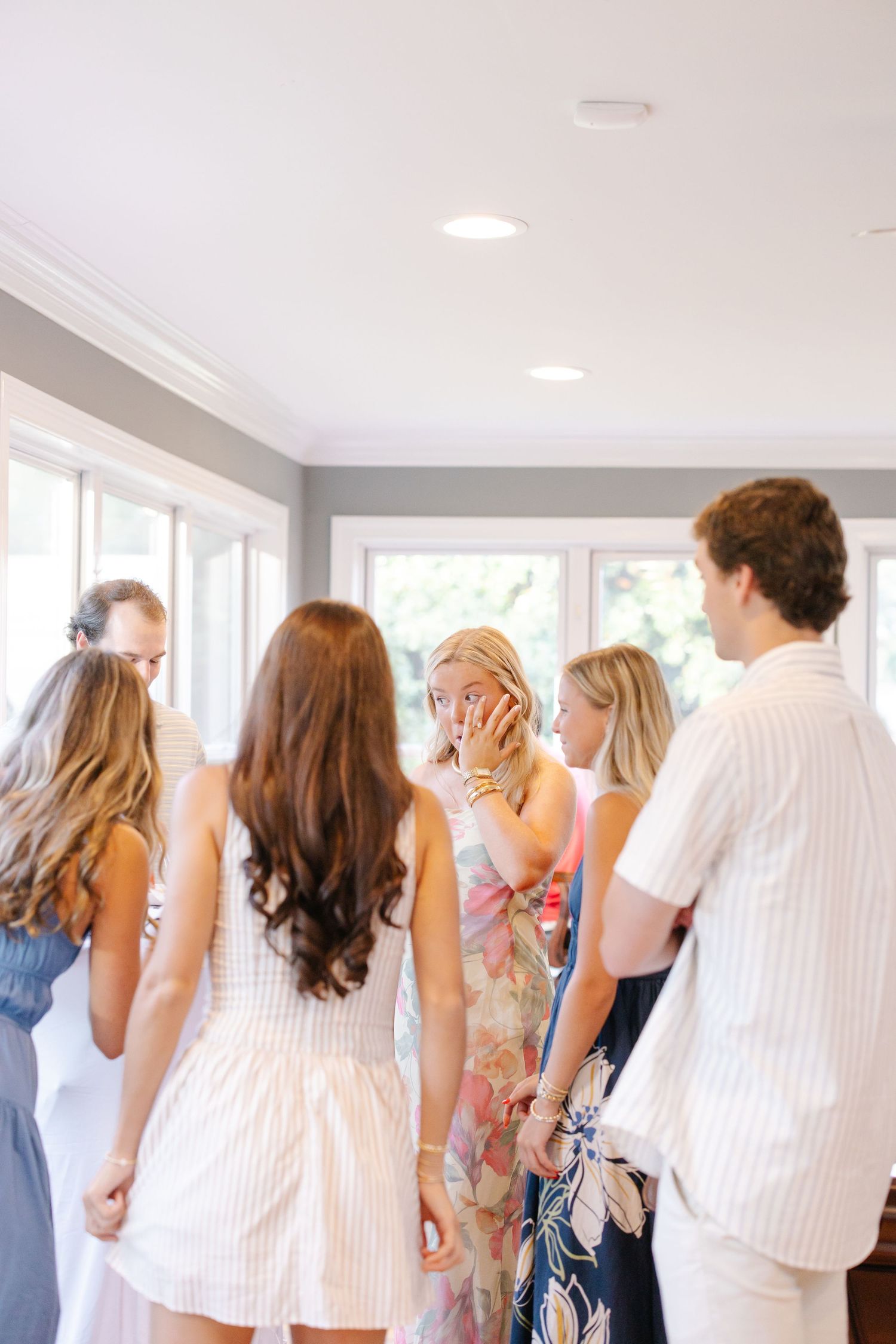 Blonde woman covering her mouth in surprise while surrounded by friends at an indoor gathering.
