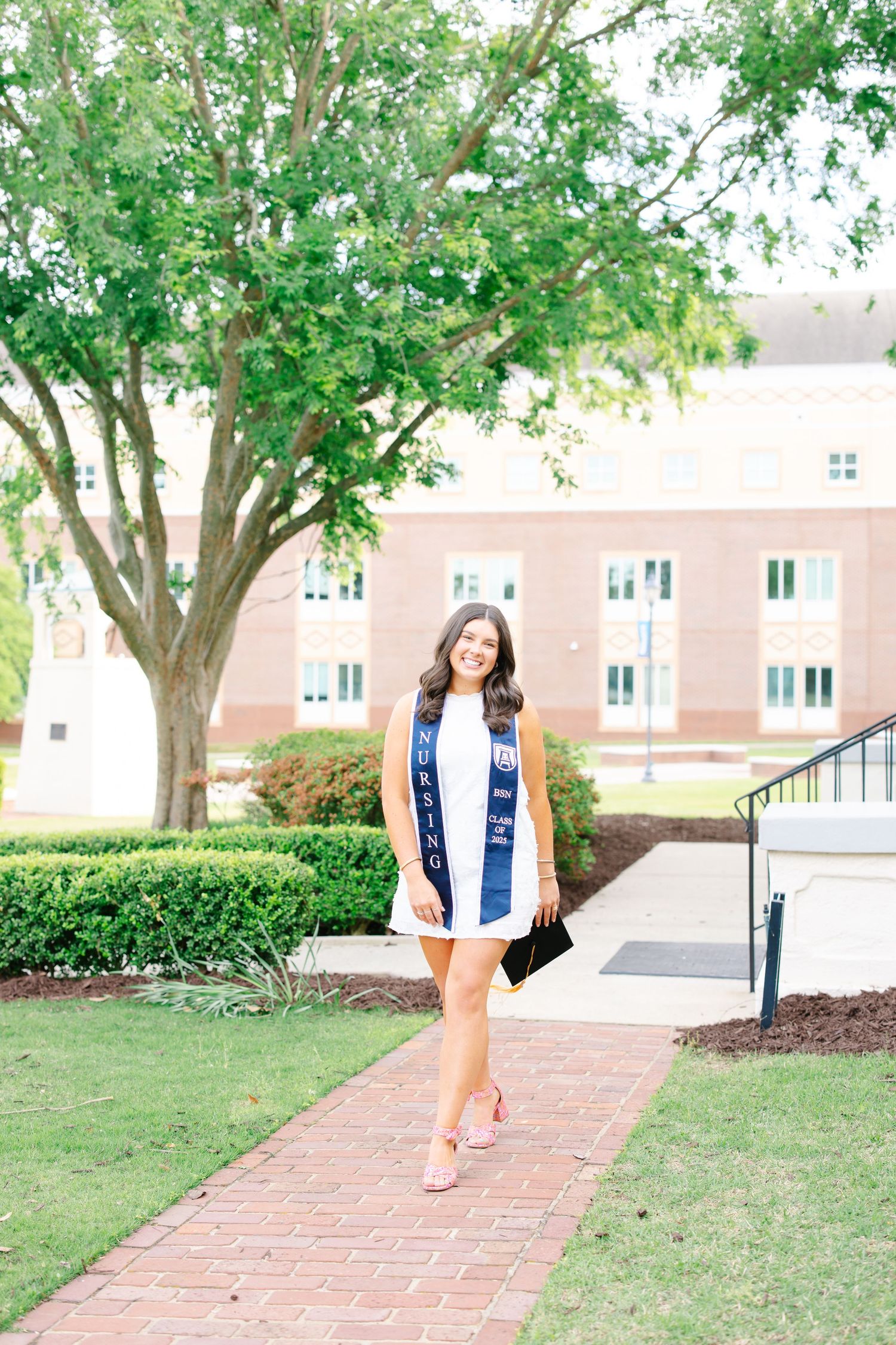 Graduating student in white dress with navy blue nursing stole walking on brick campus path.