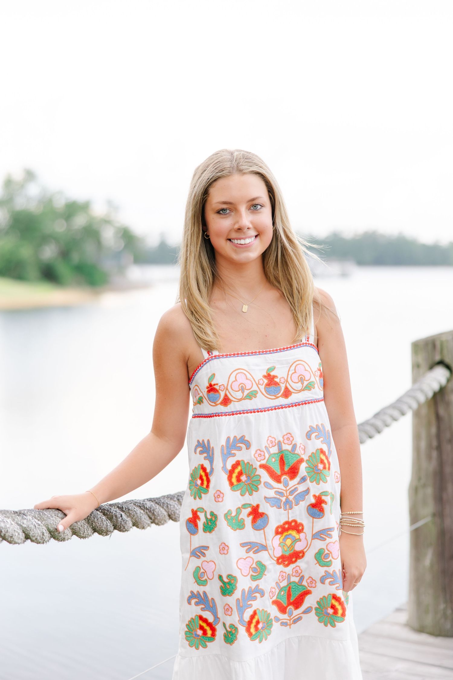 Smiling blonde teen in colorful embroidered white sundress posing on dock holding rope railing by lake.