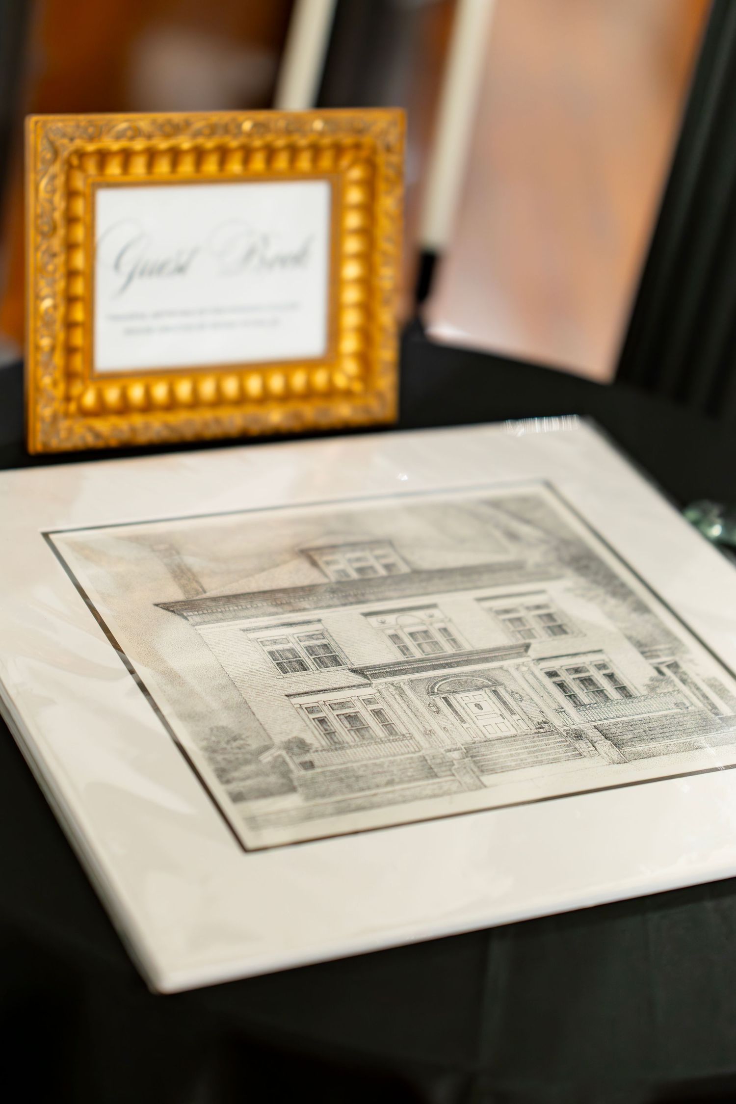 Elegant wedding guest book display with gold ornate frame sign and architectural sketch drawing on black table.