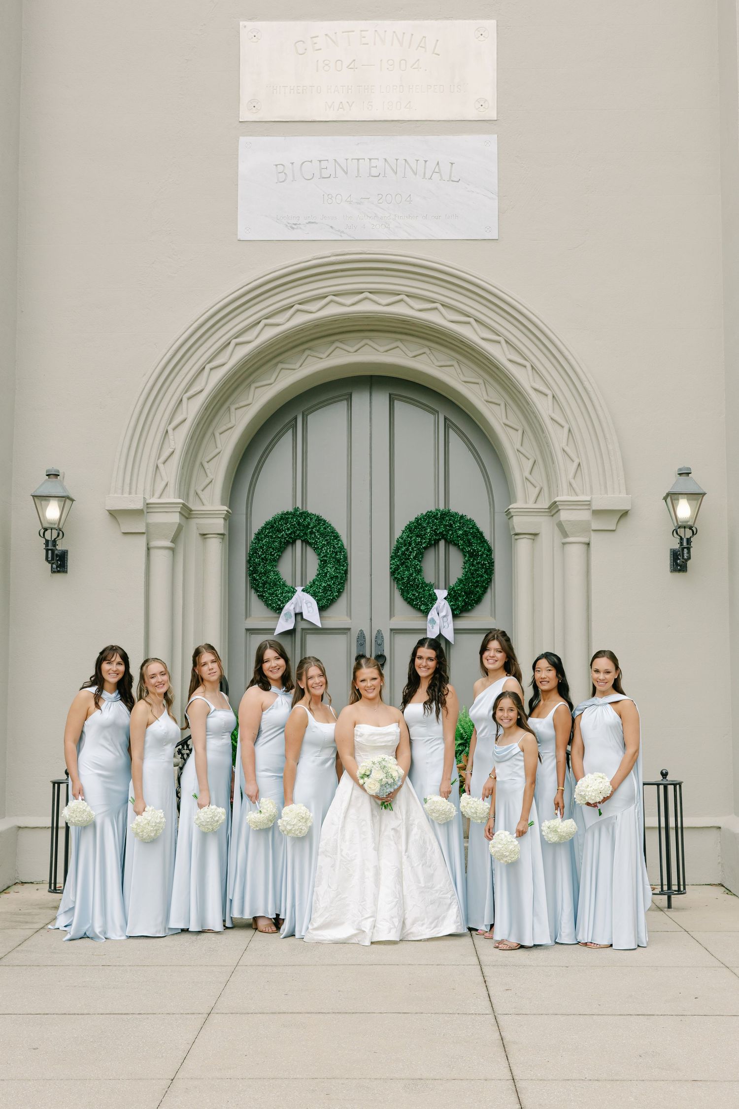 Bride in white ball gown with bridesmaids in light blue gowns holding white bouquets outside church.