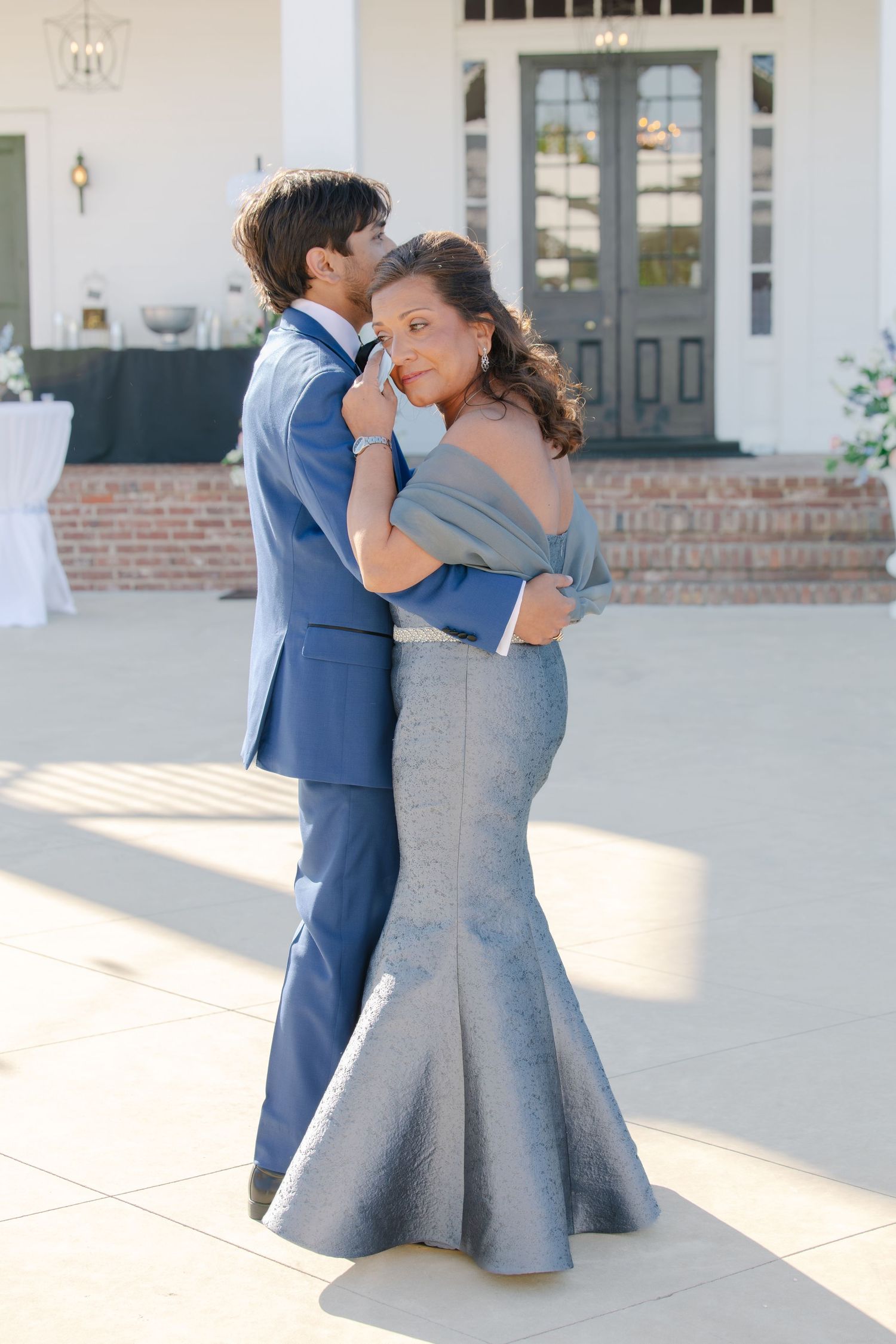 Groom in blue suit sharing a tender dance with woman in elegant grey off-shoulder mermaid gown outside white venue.