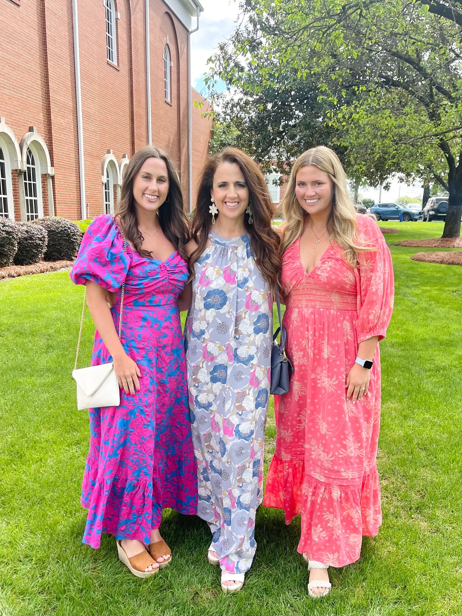 Three women in colorful floral maxi dresses posing together on a church lawn at an outdoor event.