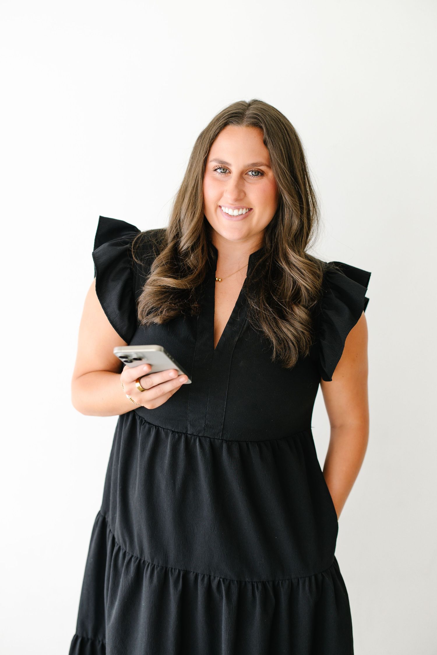 Smiling brunette woman in a black ruffled dress holds a smartphone against a white background.