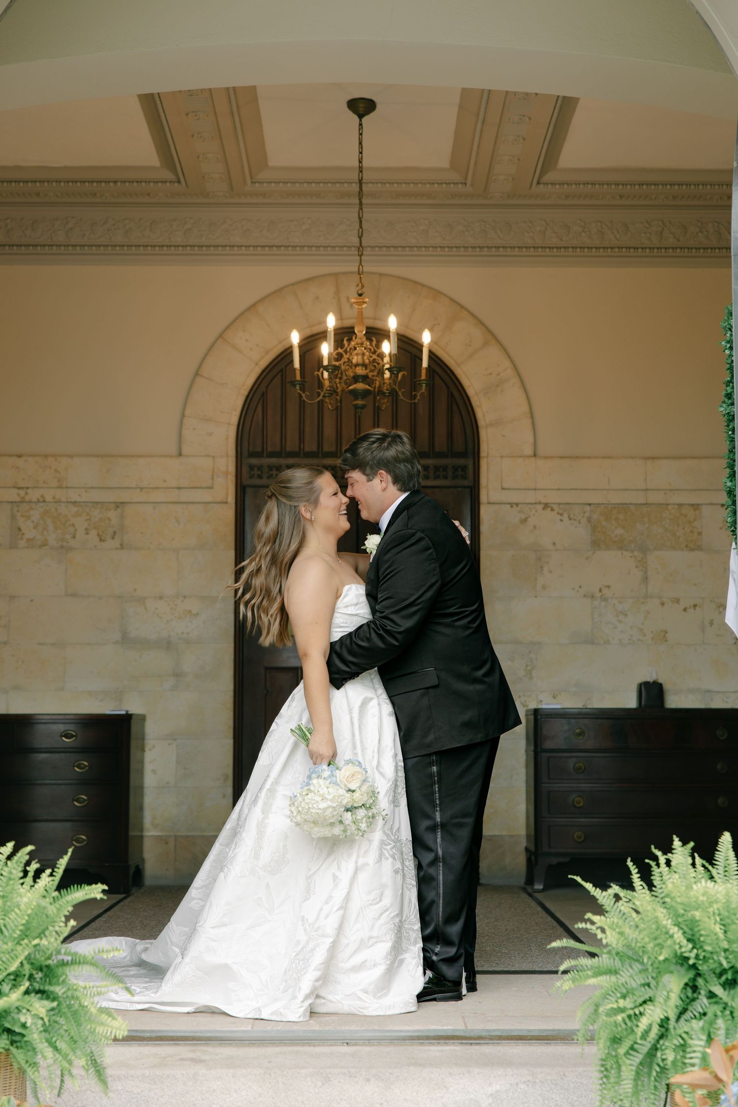 Bride and groom share first kiss under chandelier at elegant stone venue, bride holding white bouquet in strapless gown.