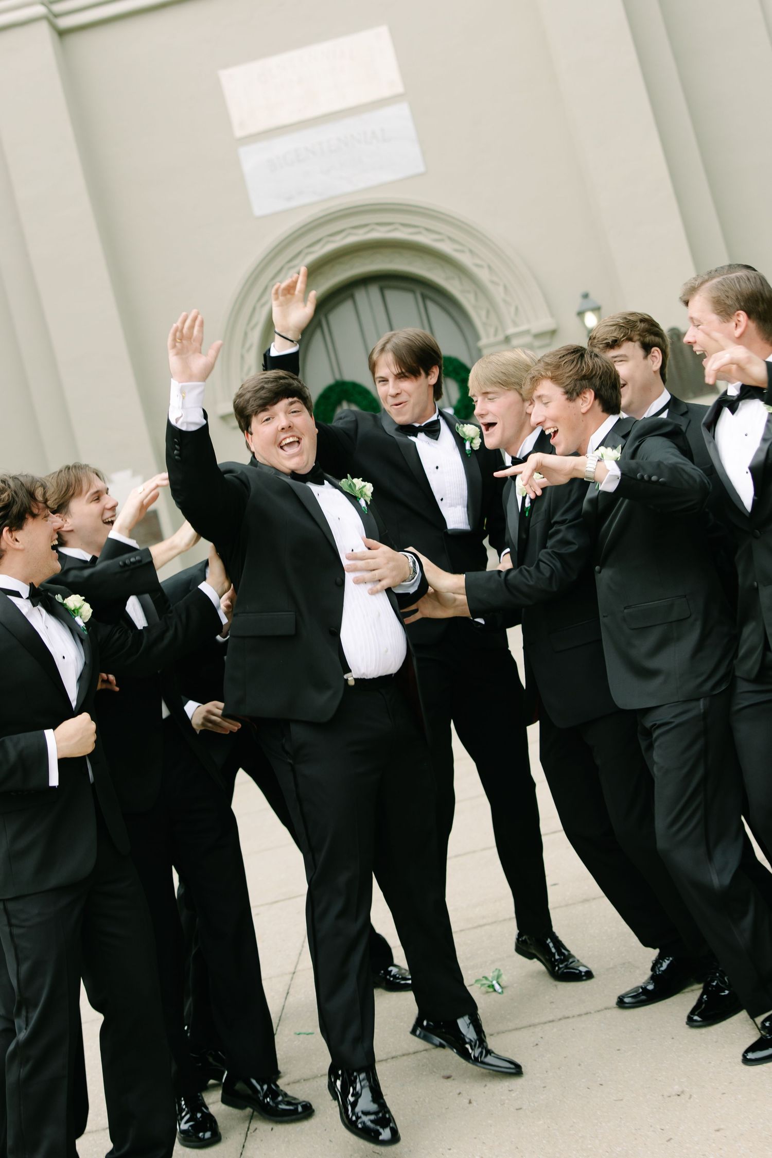 Groom and groomsmen in black tuxedos celebrate energetically outside a church in a candid fun moment.