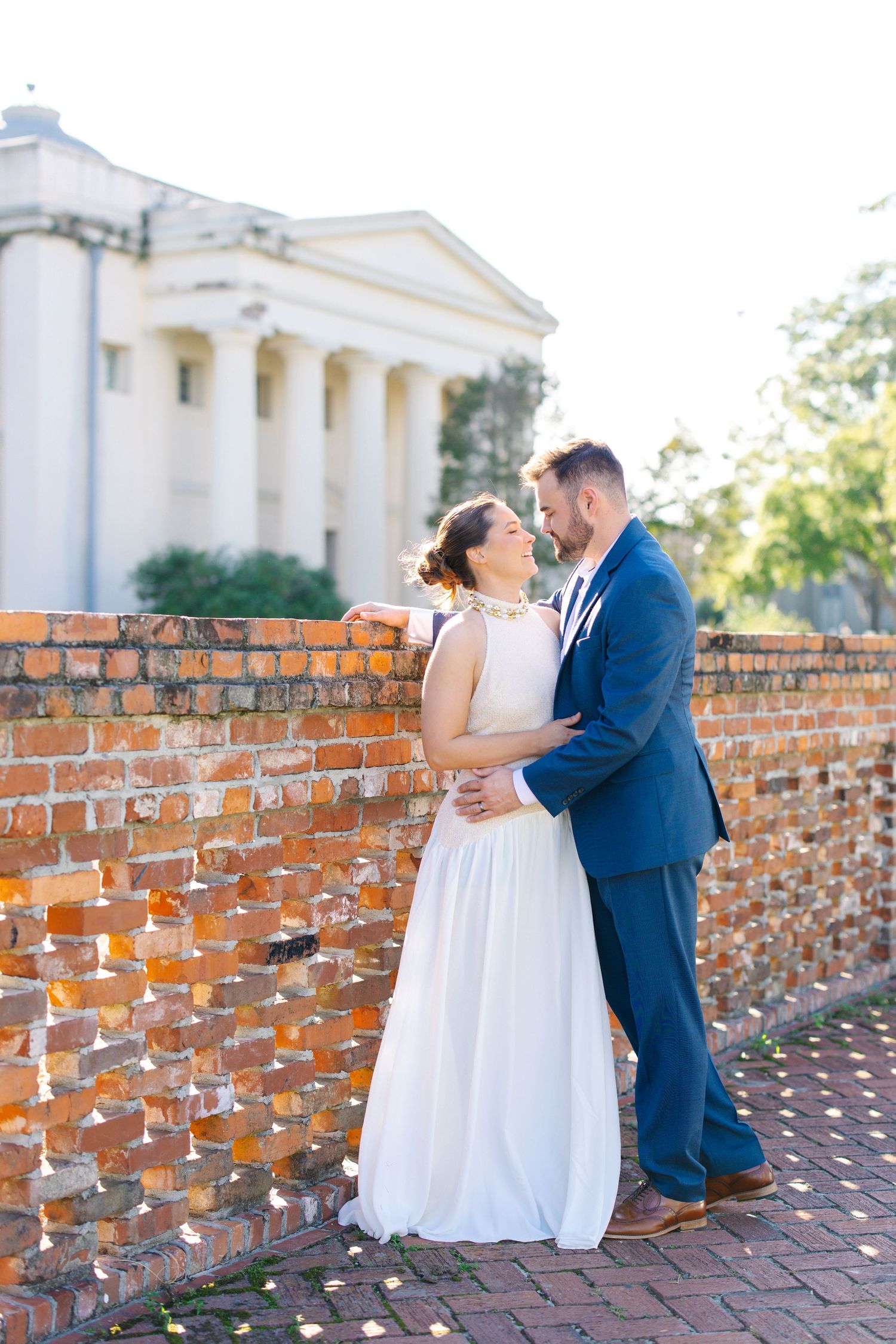 Bride in white dress and groom in blue suit embracing by rustic brick wall near white columned building.