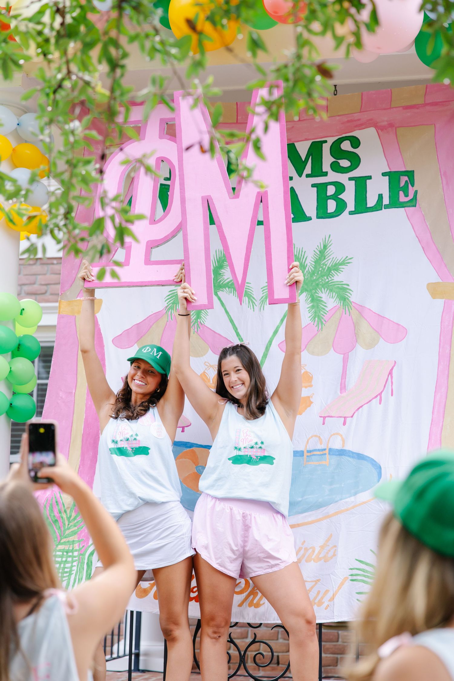 Two women hold pink letter signs at a Phi Mu sorority bid day event with tropical themed decorations.