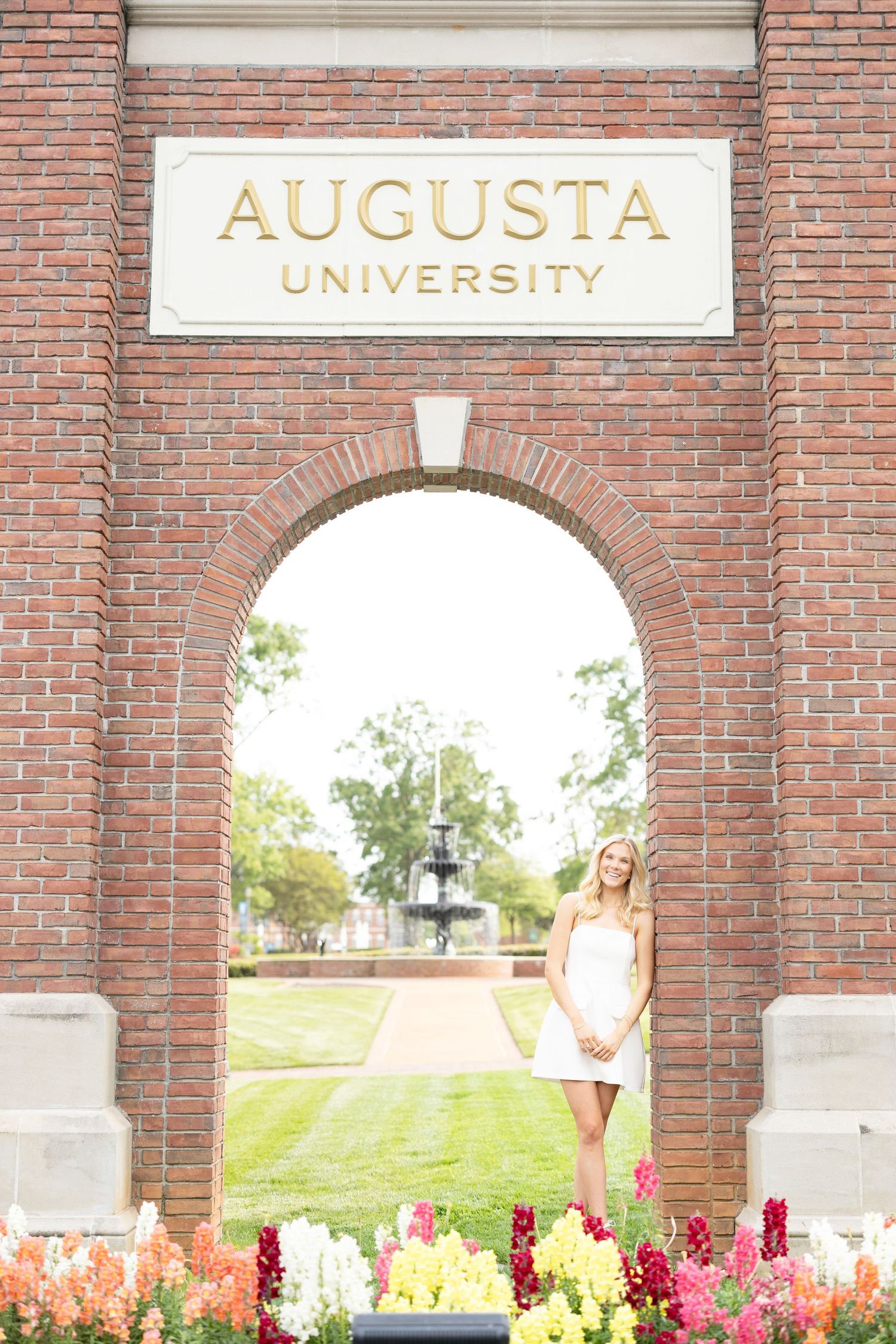 Blonde graduate in white dress standing under Augusta University brick arch with colorful flowers.