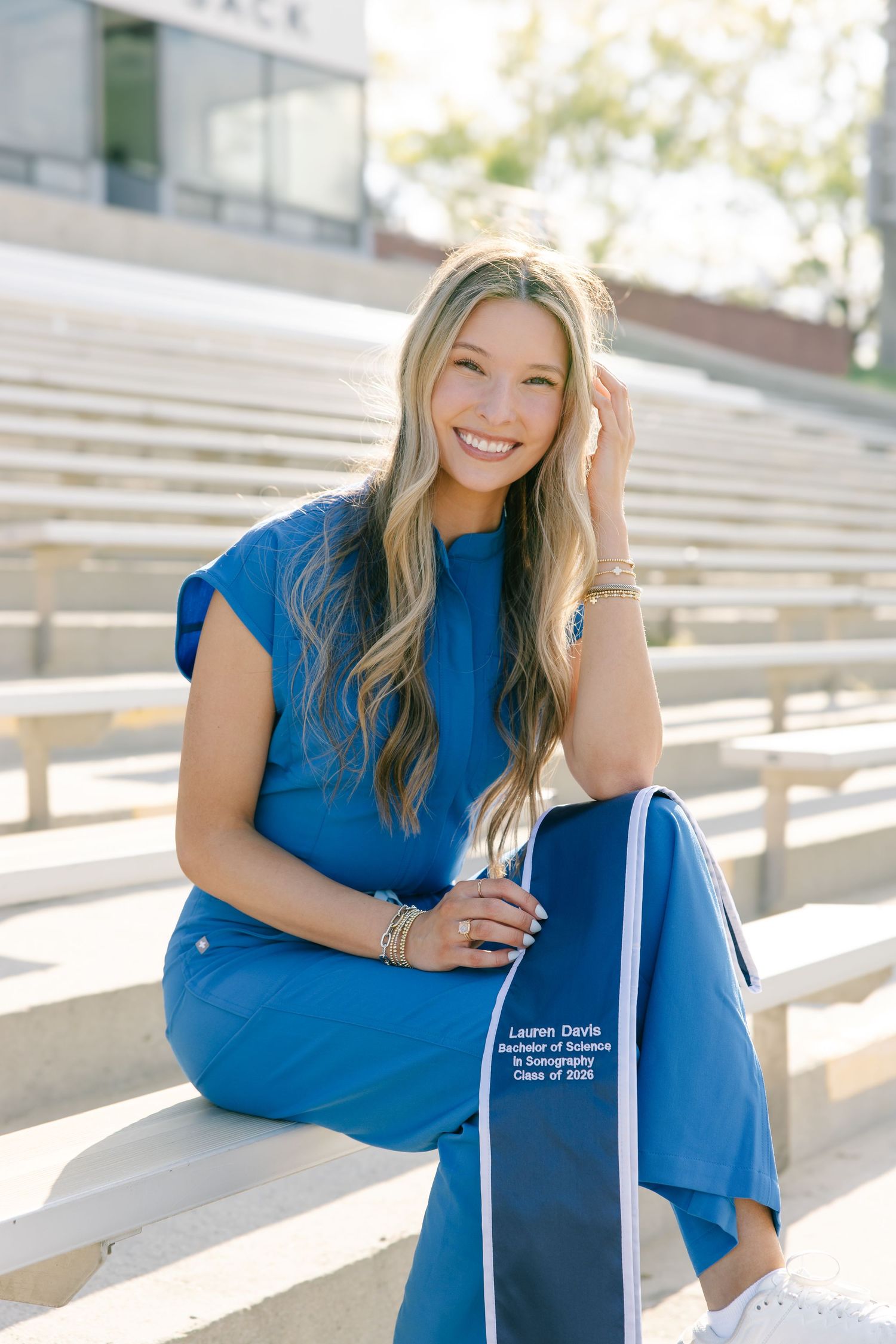 Smiling blonde graduate in blue outfit seated on stadium bleachers holding graduation stole in sunlight.