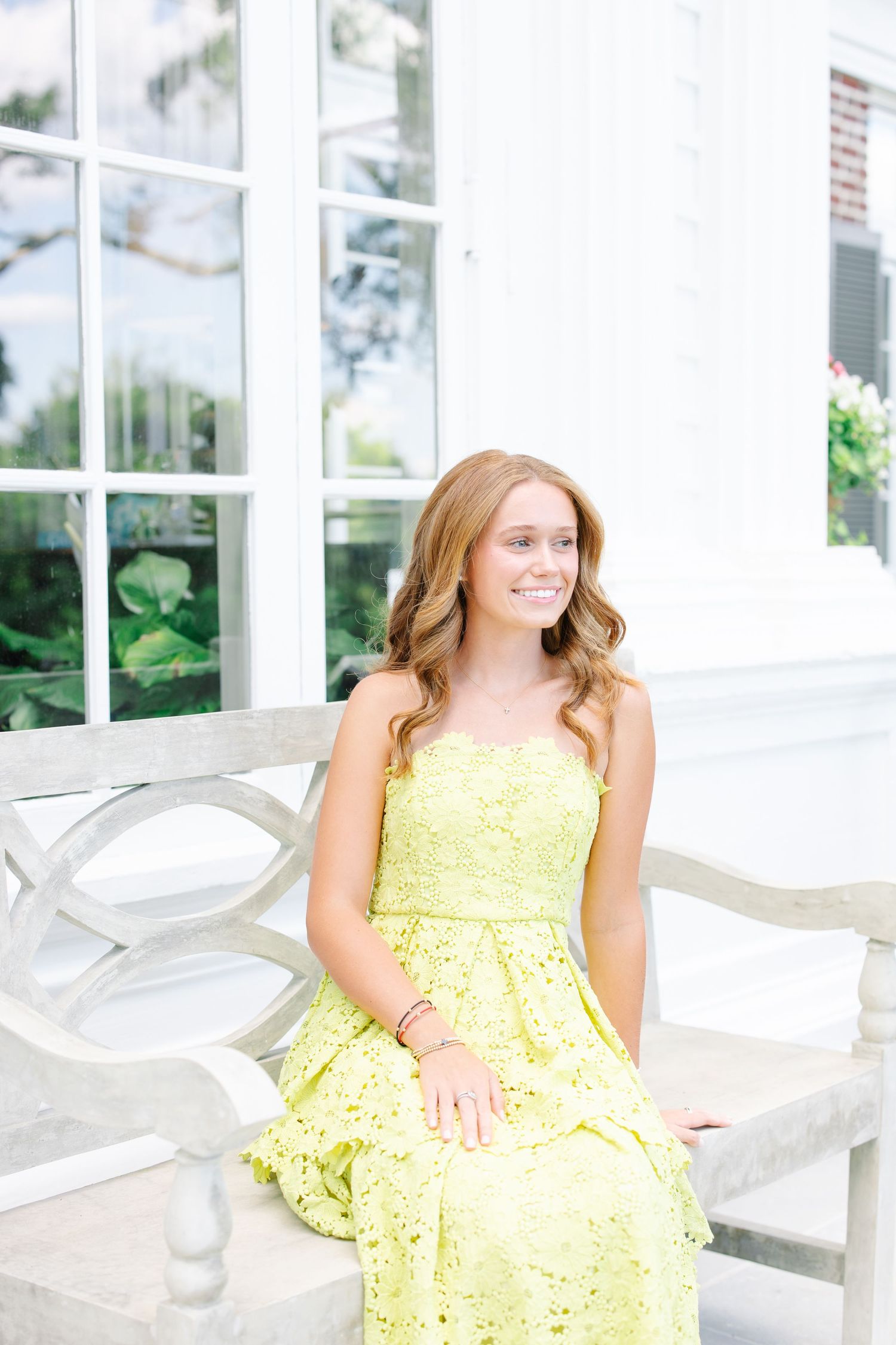 Smiling teen in yellow lace sundress sitting on a white ornate bench outside a white building.