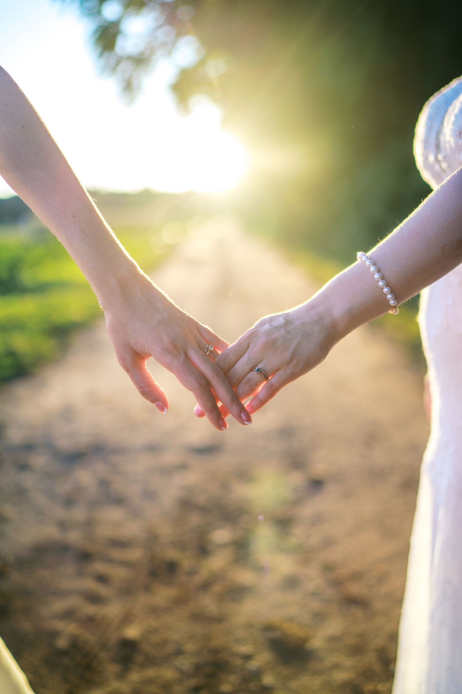 Fernbrook farms Philly wedding Two brides holding hands with wedding rings on a sunlit path, wearing white dress and pearl bracelet.