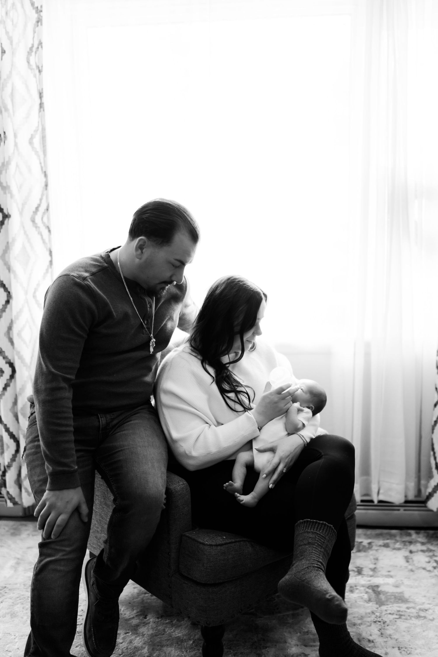 Black and white photo of parents admiring their newborn baby, mother seated in chair with father leaning in lovingly.