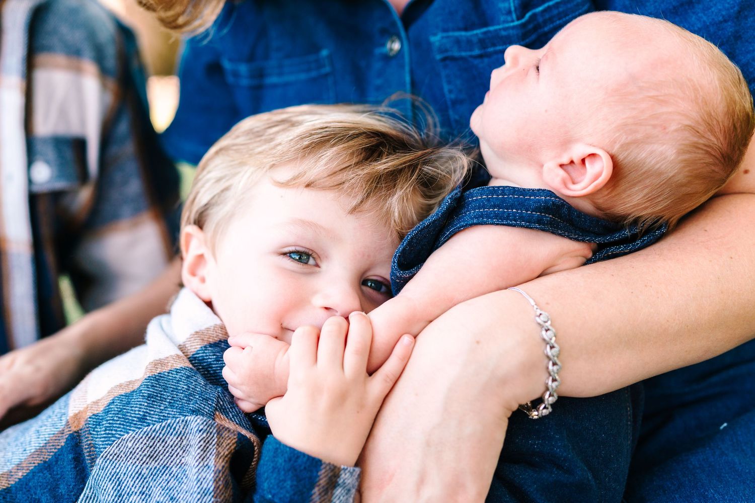 A toddler with hands near mouth smiles beside a newborn baby held by an adult in denim outdoors.