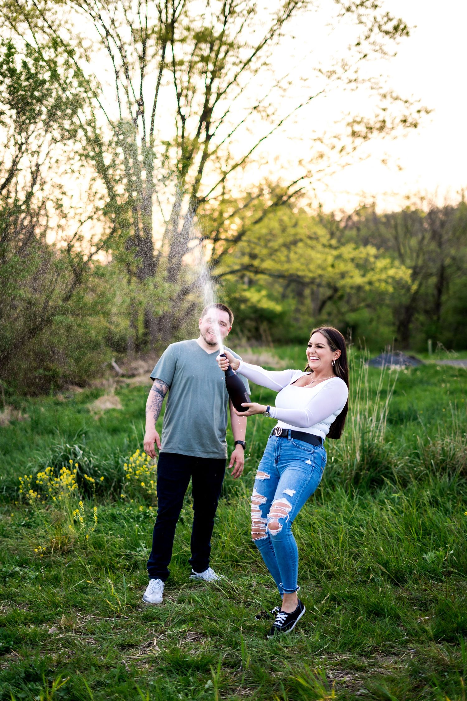 Couple posing outdoors at sunset in a green field, woman playfully holding a smoke effect behind man in gray shirt.