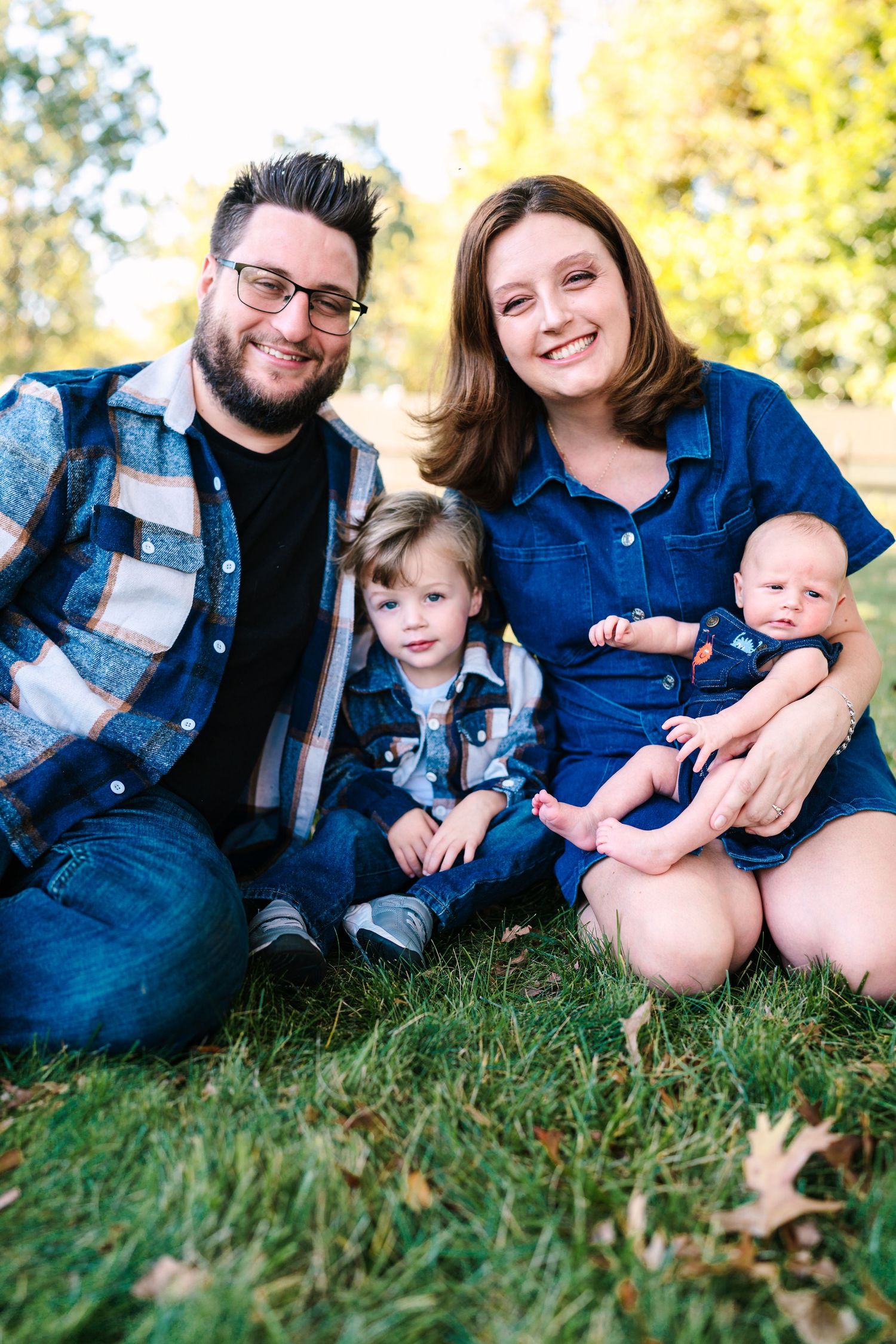 Happy family of four sitting on grass in a park, parents with toddler and newborn baby, wearing coordinating blue outfits.