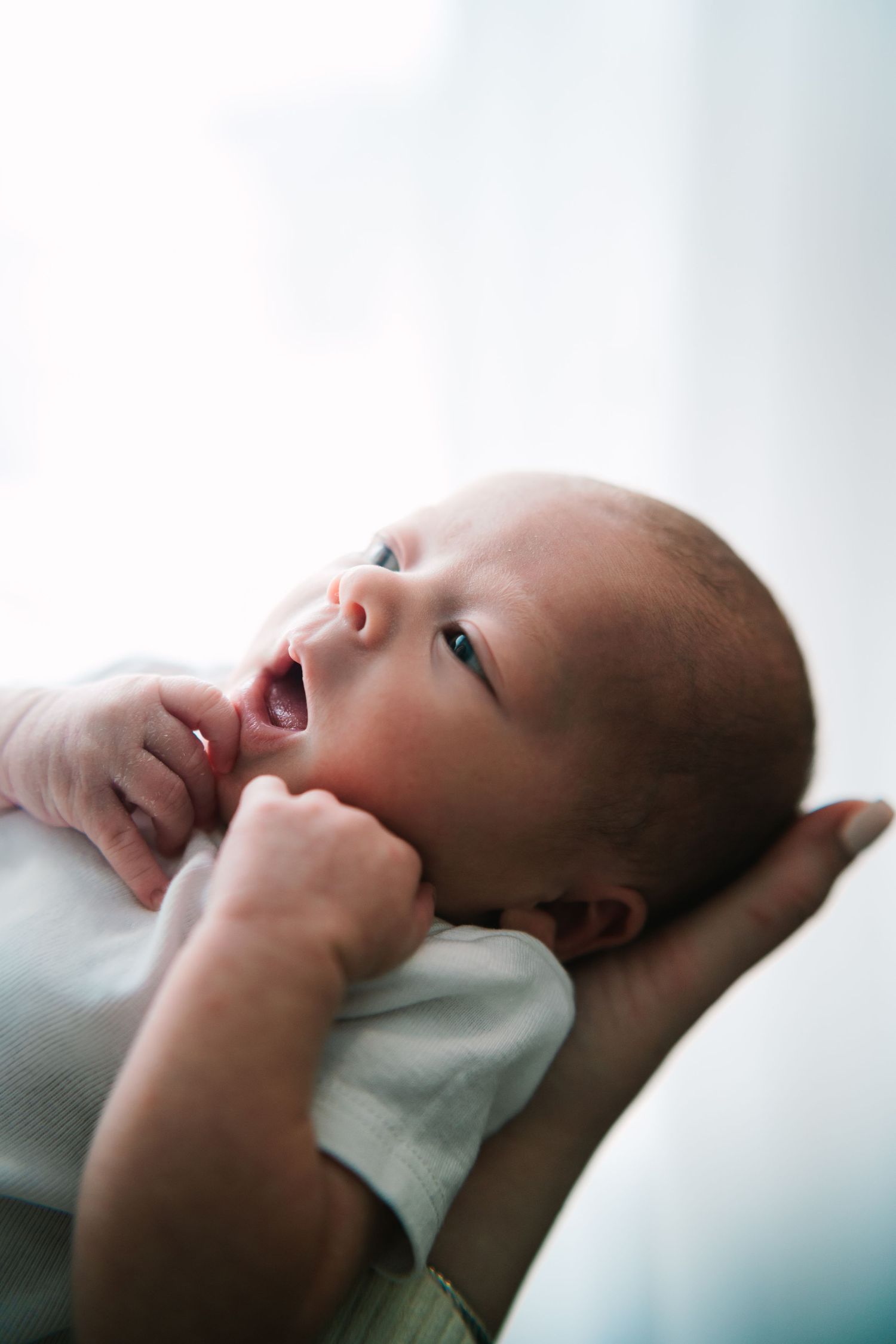 A newborn baby with mouth open being cradled in adult arms, looking upward in soft natural light.