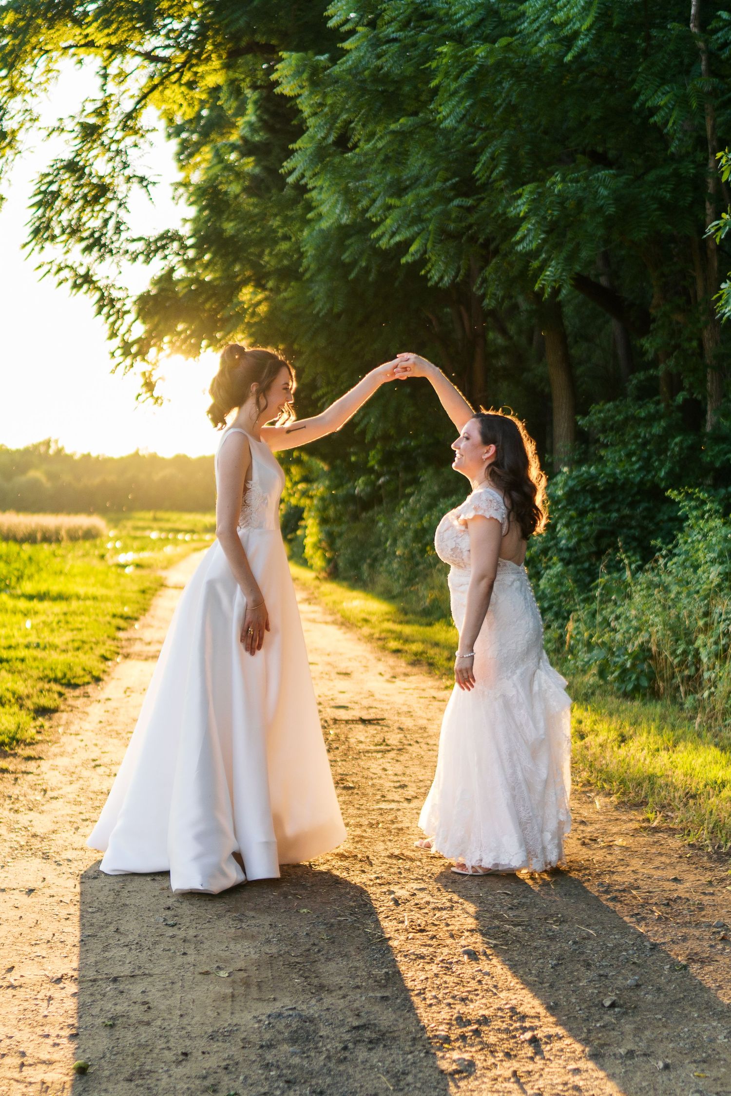 Fernbrook farms Philly wedding photo brides in white gowns hold hands on a sunlit path surrounded by lush green trees during golden hour.