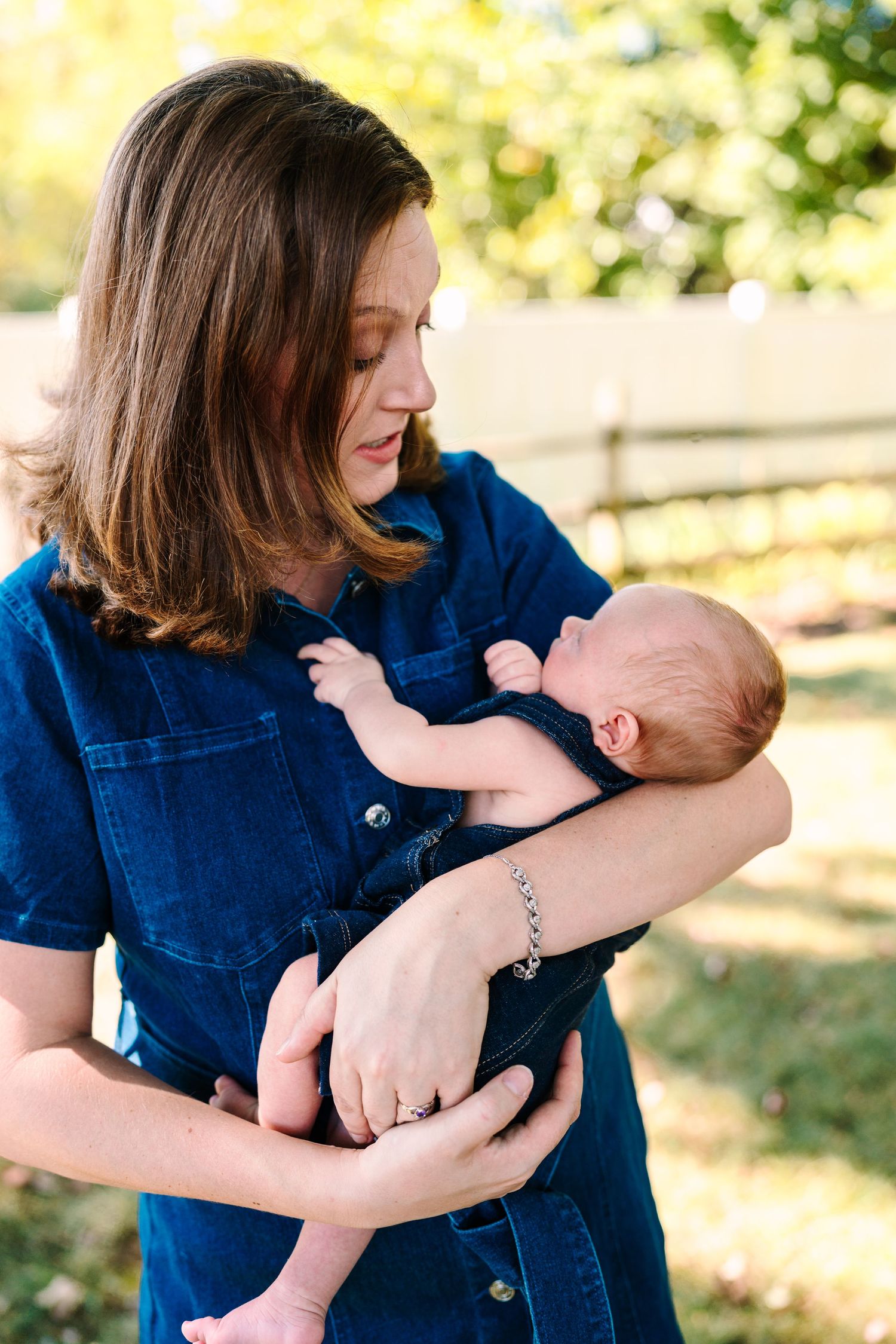 A young mother in a denim dress tenderly holds her newborn baby outdoors in a sunlit garden setting.