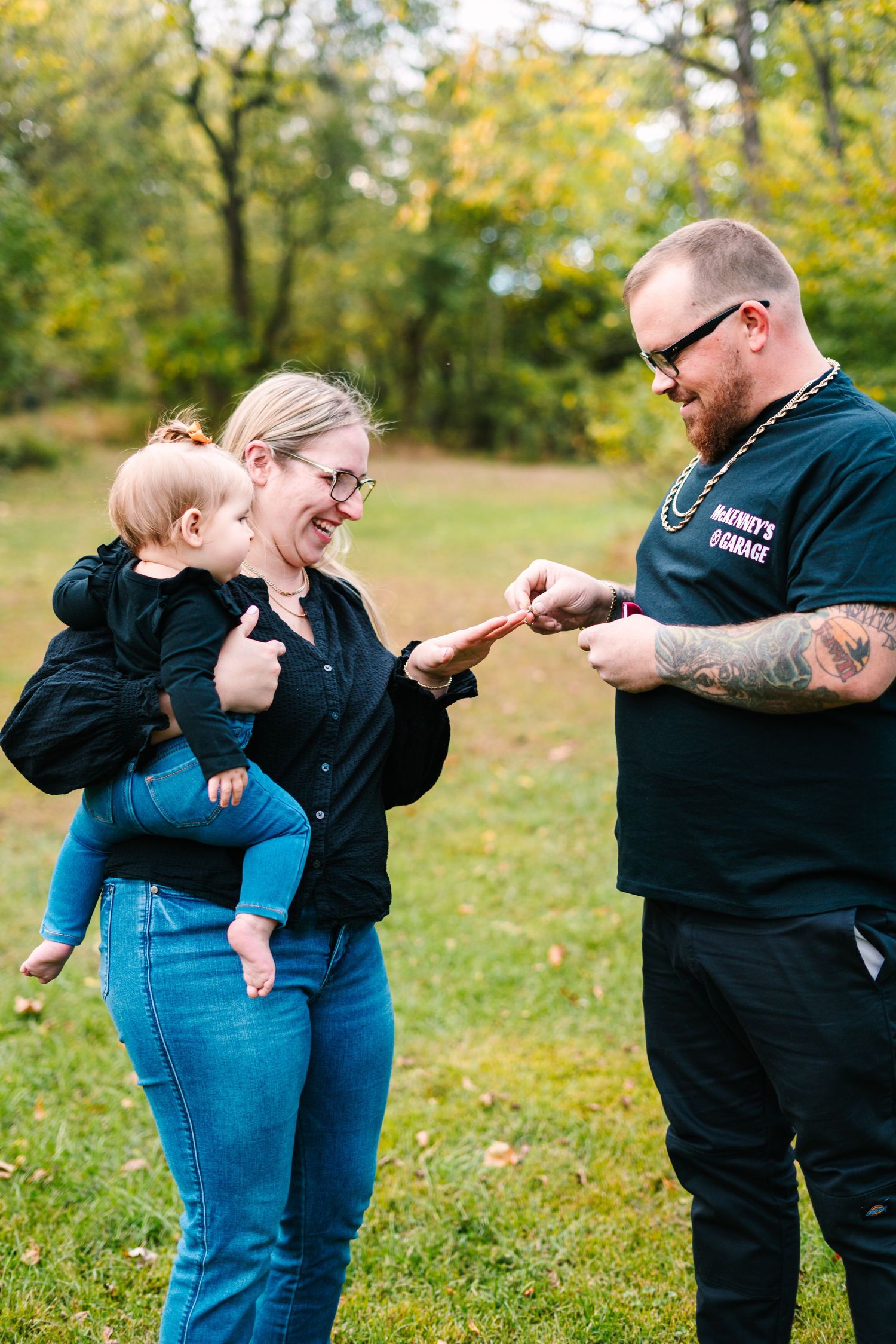 philadelphia wedding photoA couple smiles at their baby outdoors in a green park during autumn, with the man showing something to the child.