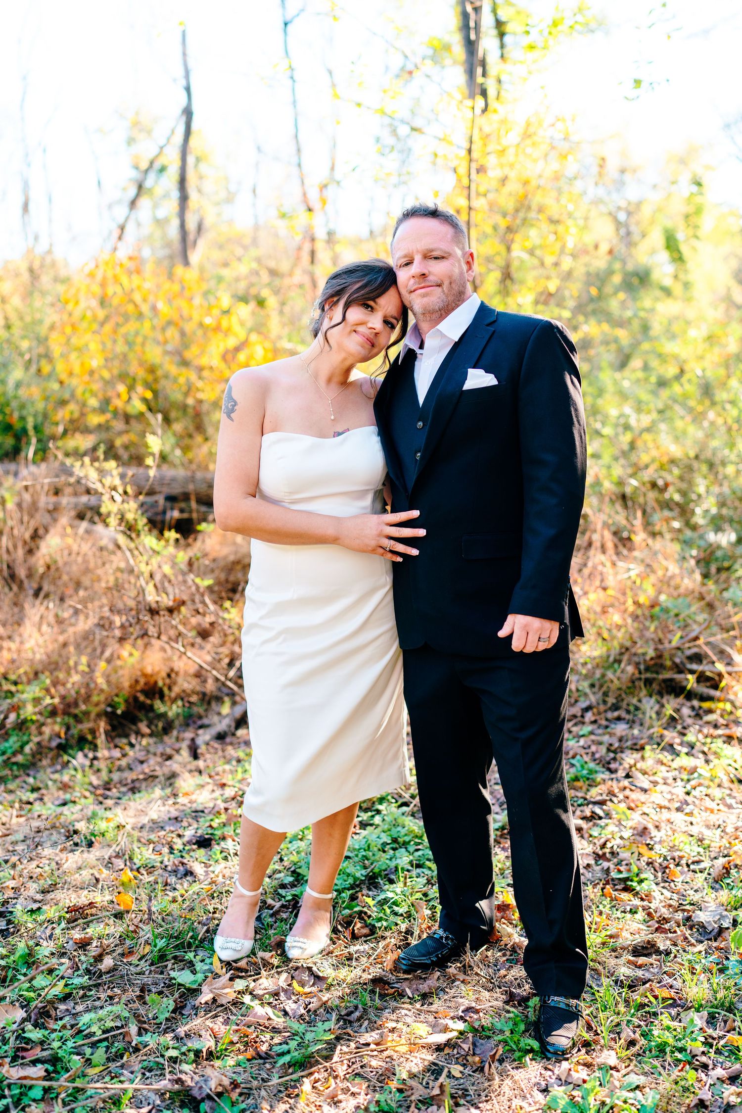philadelphia wedding photography  Bride and groom pose together in autumn woodland setting, she in white midi dress, he in black suit.