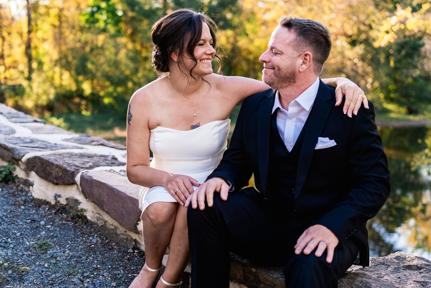philadelphia wedding Happy couple in wedding attire sitting together outdoors near a stone wall with autumn foliage backdrop.