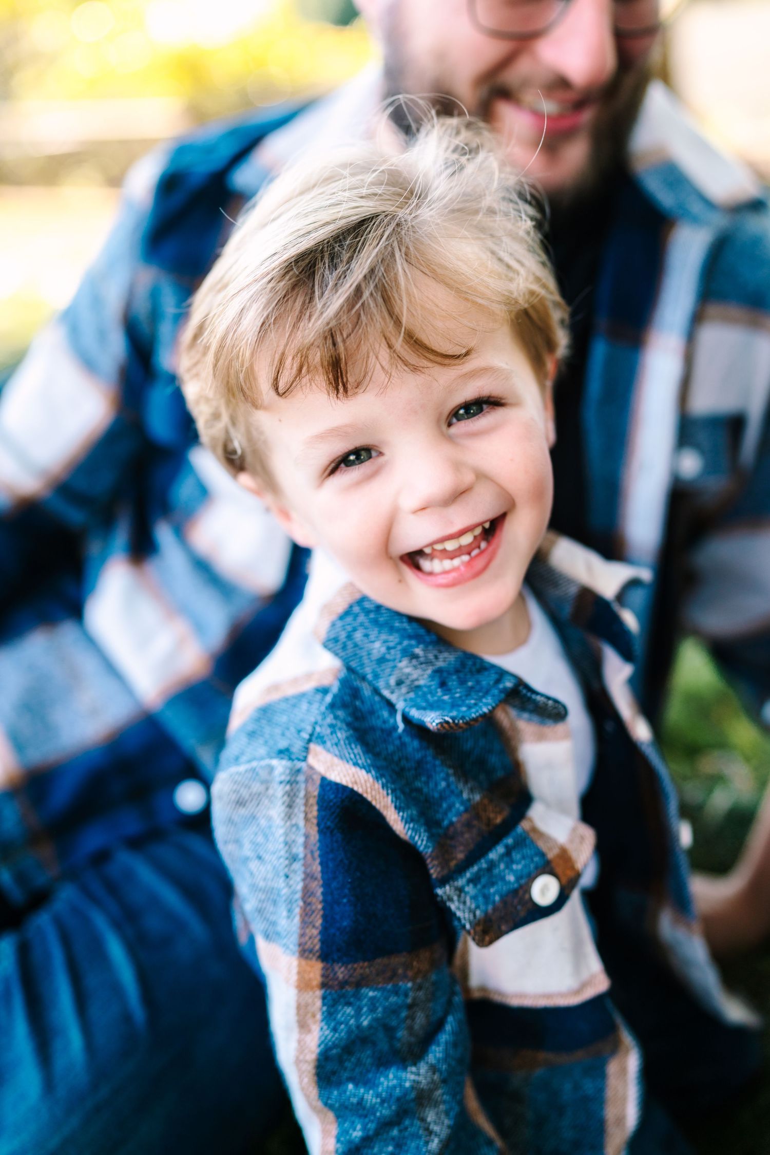 A joyful young boy in a plaid shirt laughs outdoors, with a smiling man in a matching plaid shirt behind him.